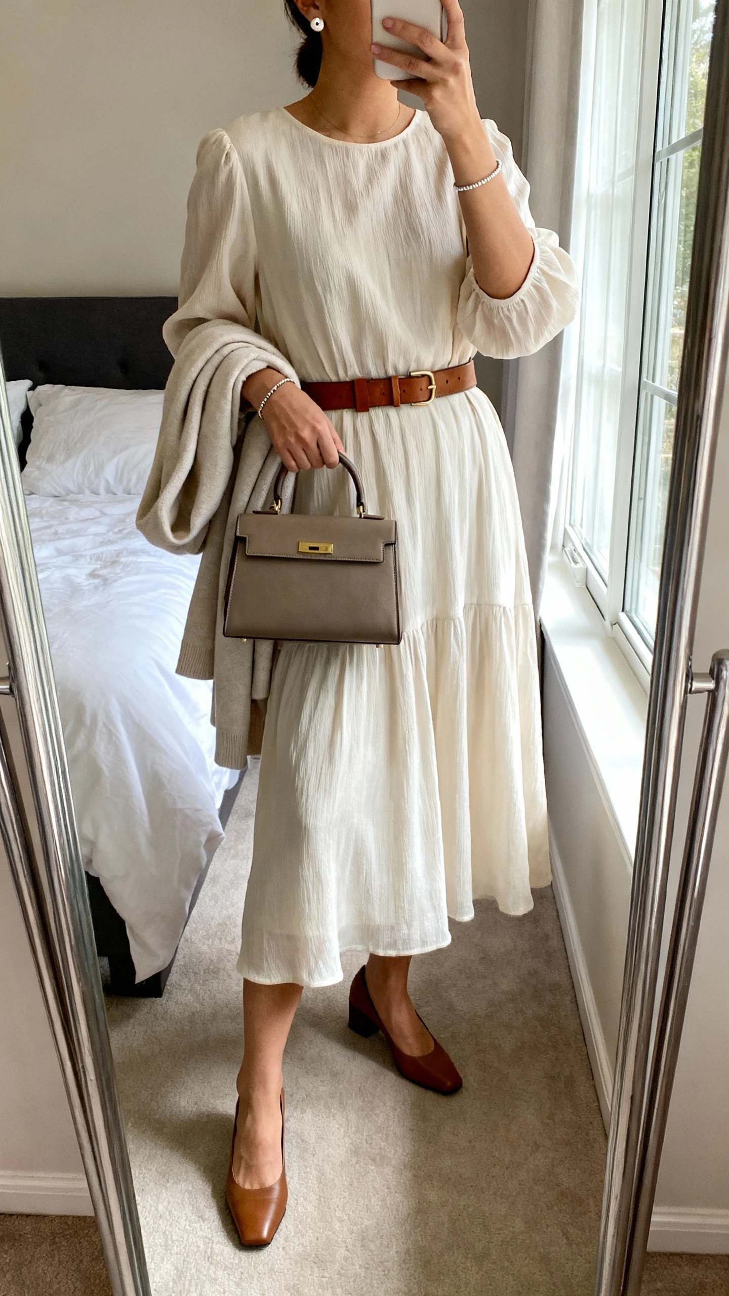 iPhone mirror selfie of a woman wearing a flowy midi dress with sleeves, a classic leather belt, low block-heel pumps, delicate stud earrings, a simple bracelet, a structured top-handle bag, and a light cardigan draped over her arm, face not visible, cropped at neck, bedroom mirror, natural window light, iPhone photo quality.