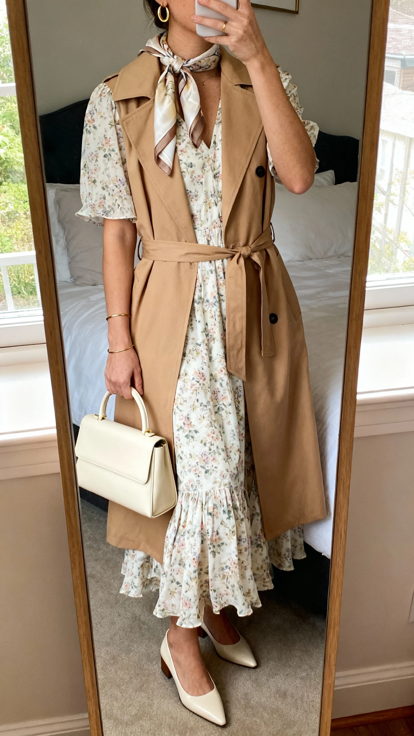 iPhone mirror selfie of a woman wearing a flowy midi floral dress with short sleeves, a lightweight belted trench coat in camel, pointed low block heels, a structured cream top-handle bag, delicate gold hoop earrings, a thin bracelet, and an optional silk scarf draped at the neckline; face not visible, cropped at neck, bedroom mirror, natural window light, iPhone photo quality.