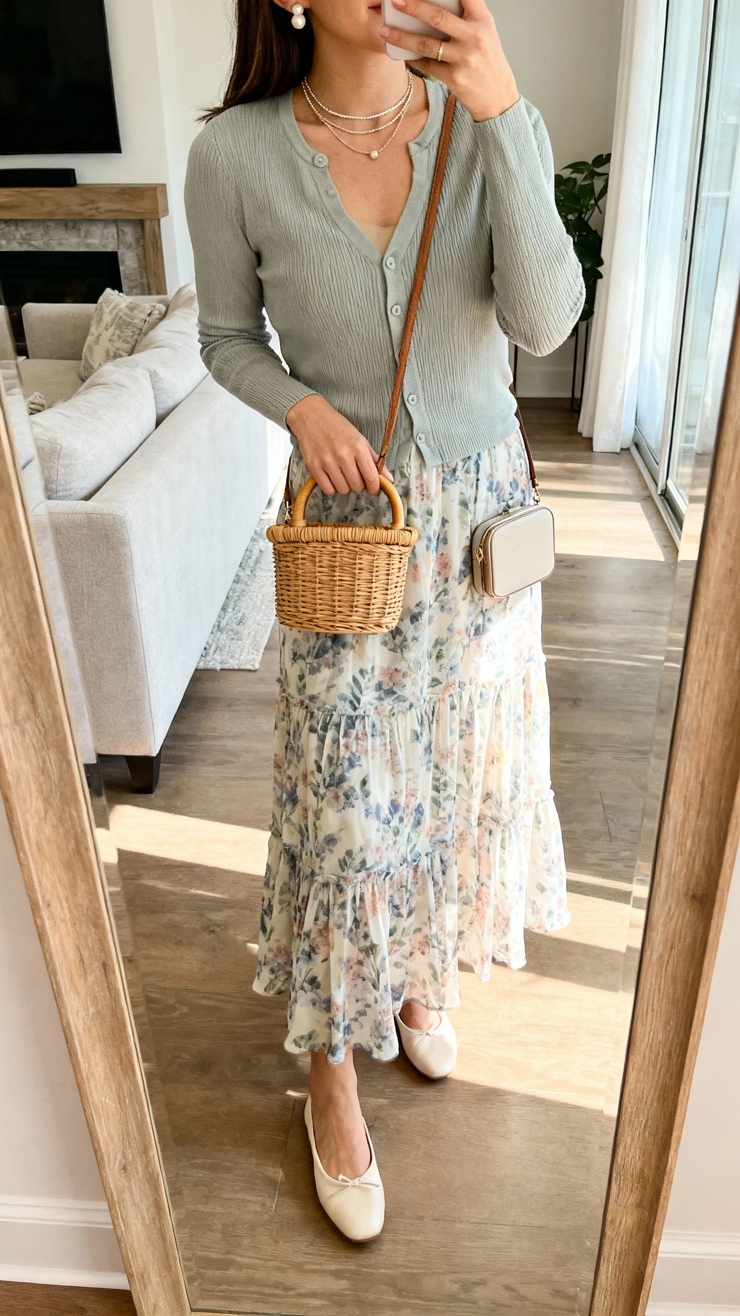 iPhone mirror selfie of a woman wearing a flowy printed maxi skirt with a fitted cardigan, sleek ballet flats, a delicate layered necklace, pearl studs, and a compact crossbody (wicker top-handle in warm months), face not visible, cropped at neck, living room full-length mirror, soft afternoon light, iPhone photo quality.