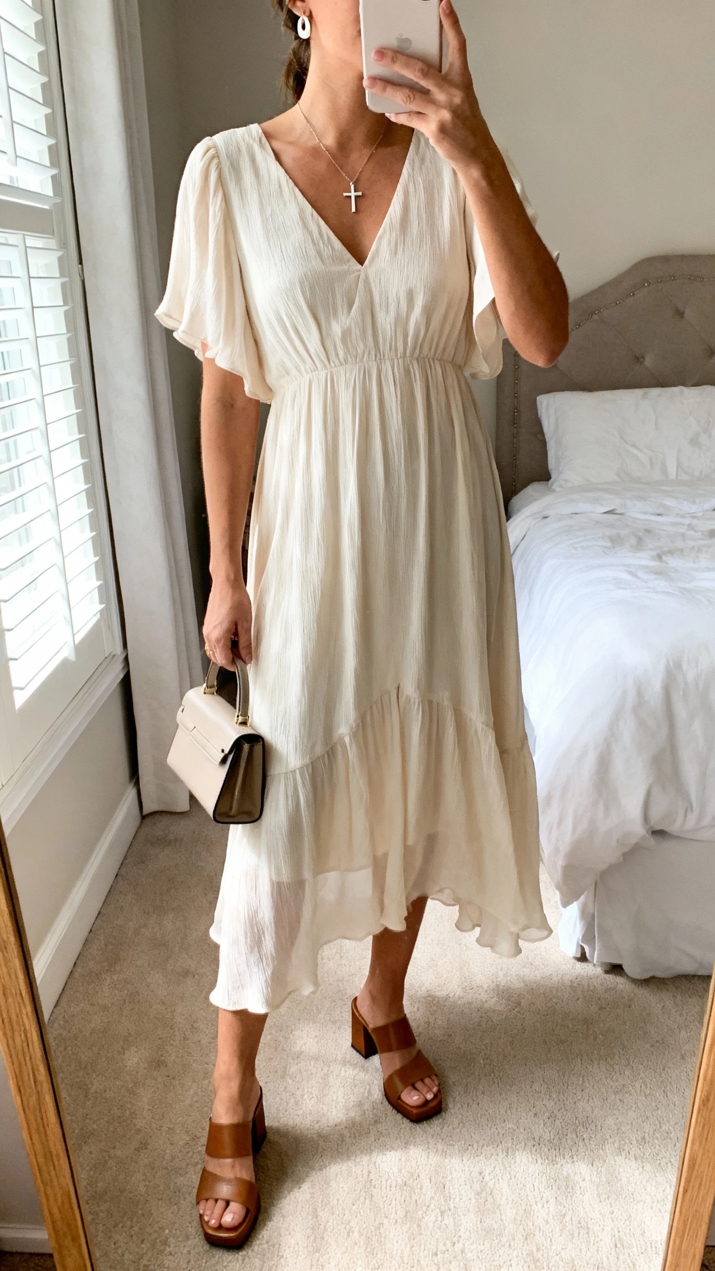 iPhone mirror selfie of a woman wearing a flowy Sunday-ready midi dress with gentle movement, delicate earrings, simple cross necklace, low block-heel sandals, and a small structured handbag, face not visible, cropped at neck, bedroom mirror with natural morning window light, iPhone photo quality.