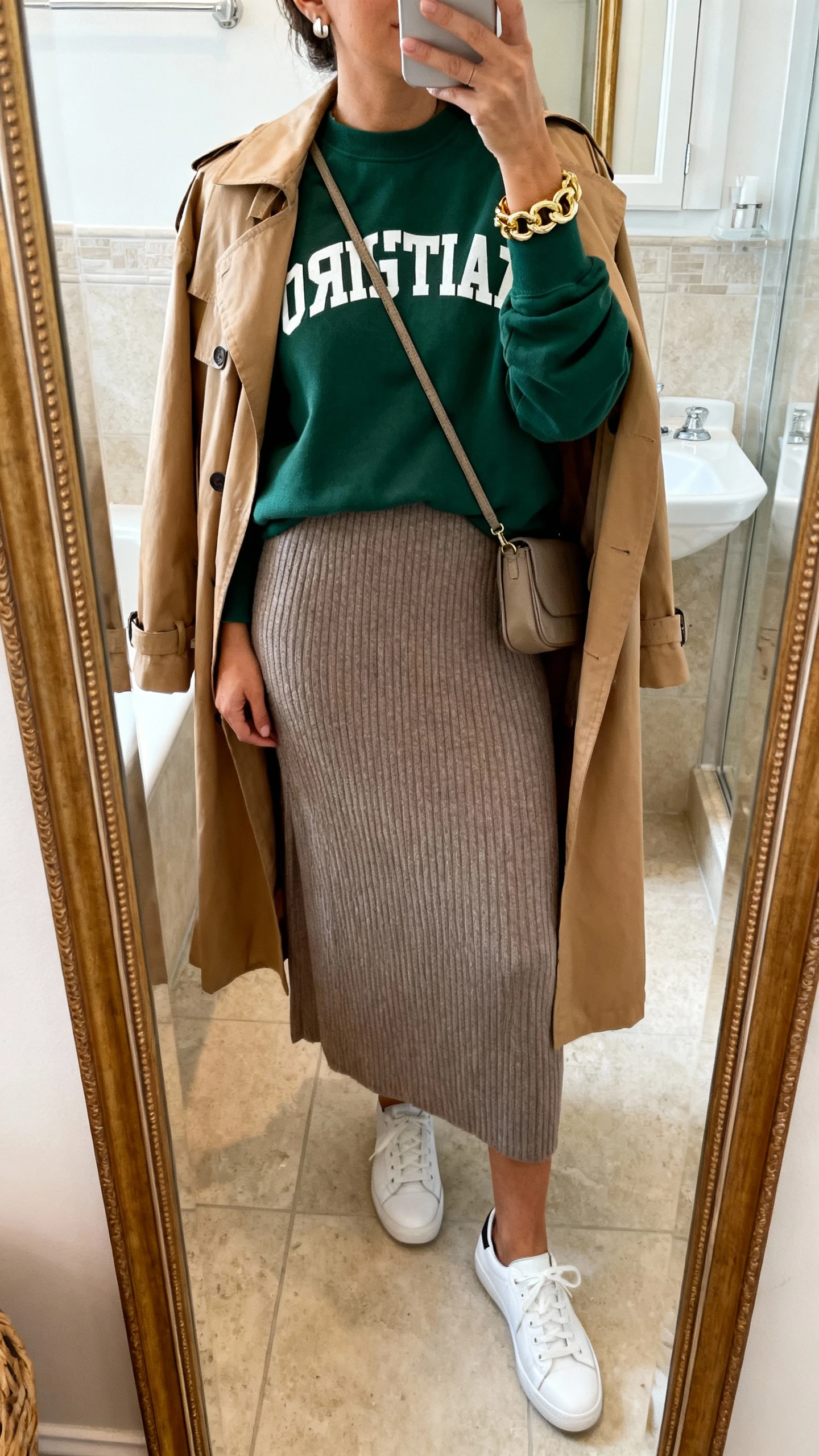 iPhone mirror selfie of a woman wearing a forest green Christian crewneck layered over a taupe ribbed knit maxi dress, classic white leather sneakers, a camel lightweight trench coat, a chunky gold chain bracelet, simple stud earrings, and a small crossbody, face not visible, bathroom full-length mirror, indoor natural light, iPhone photo quality.