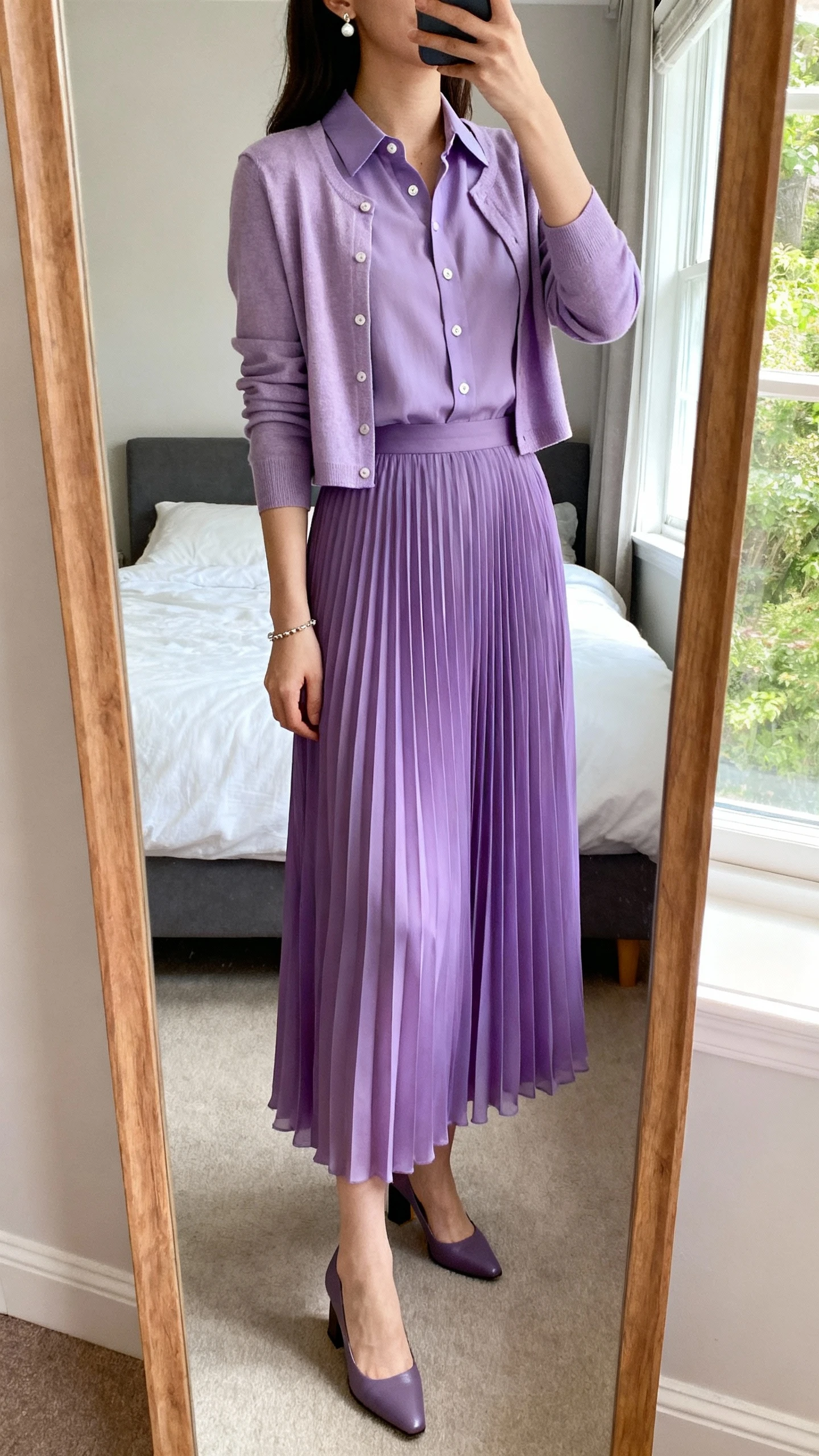iPhone mirror selfie of a woman wearing a full-length pleated chiffon skirt in a soft pastel or jewel tone, a tailored long-sleeve button-front blouse with a subtle collar, a lightweight cropped cardigan, closed-toe block-heel pumps, delicate stud earrings, and a thin bracelet, face not visible, cropped at neck, bedroom mirror, natural window light, iPhone photo quality.
