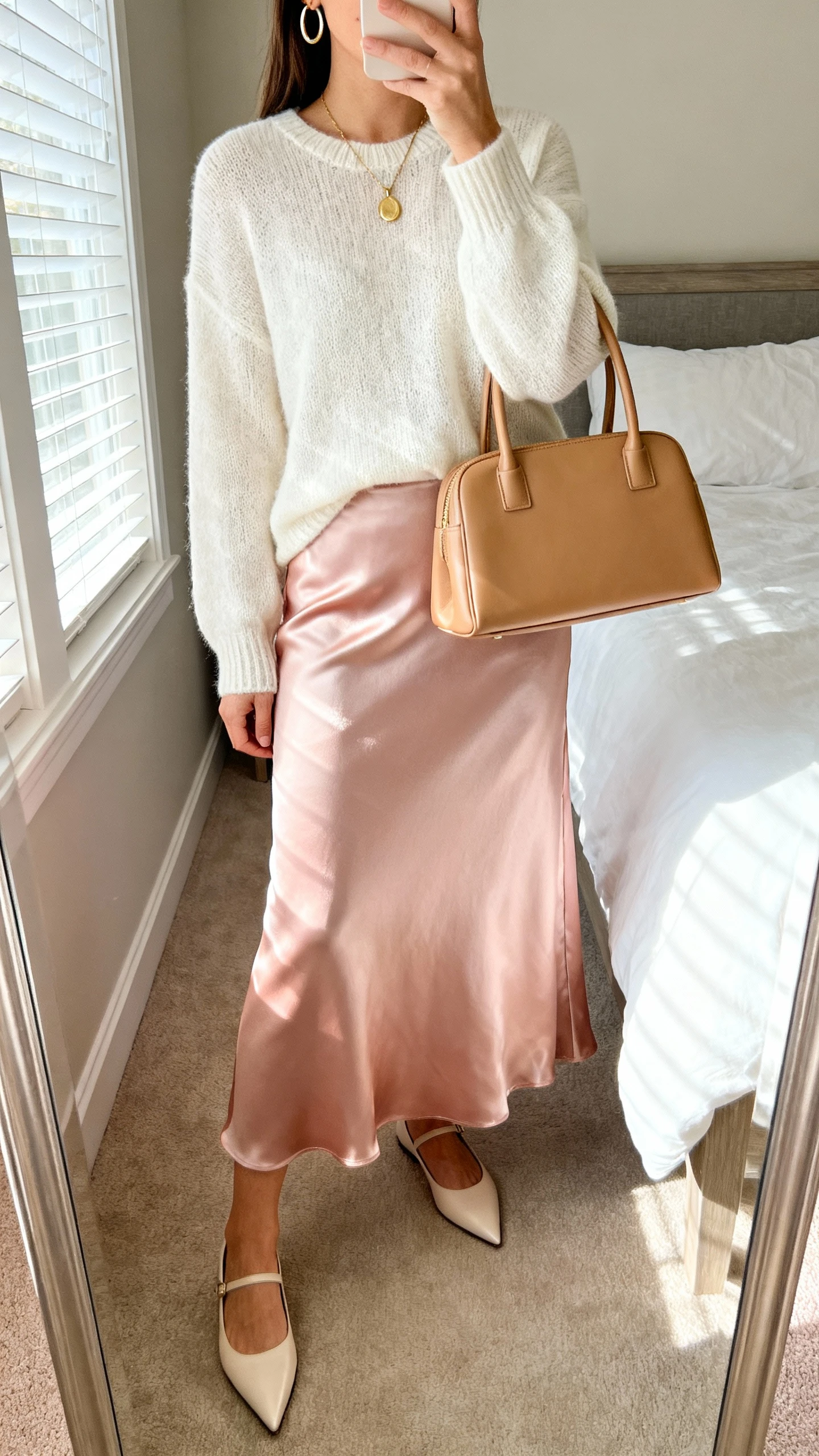 iPhone mirror selfie of a woman wearing a full-length satin skirt in blush with an ivory lightweight knit sweater, pointed-toe flats, delicate gold pendant necklace, simple hoop earrings, and a tan structured top-handle bag, face not visible, cropped at neck, bedroom mirror, soft morning window light, iPhone photo quality.