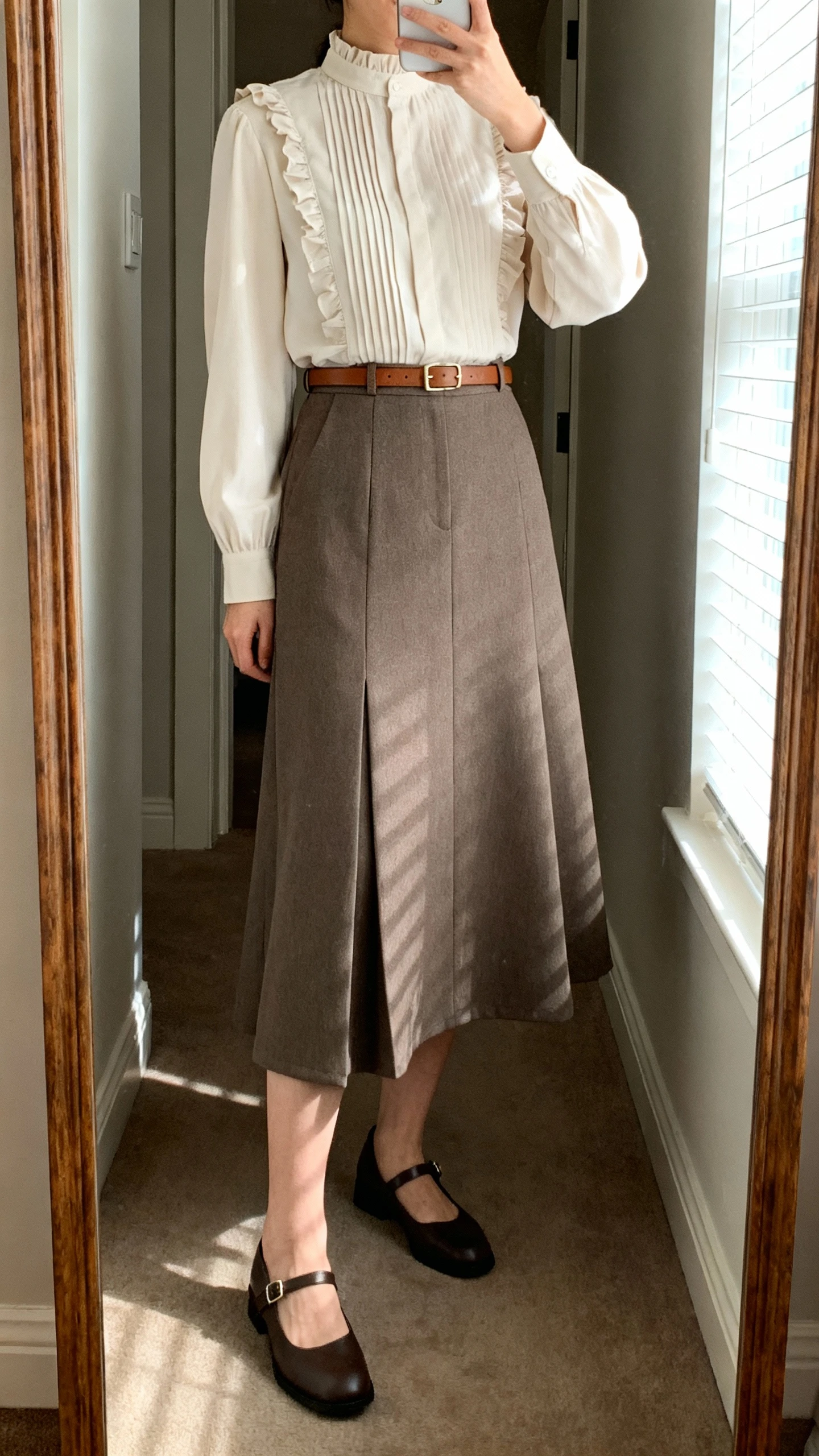 iPhone mirror selfie of a woman wearing a full-length A-line skirt in structured fabric, a modest high-neck blouse with subtle pintucks/ruffles, a slim leather belt, and classic Mary Janes, face not visible, cropped at shoulders, full-length mirror in hallway, morning window light, iPhone photo quality.