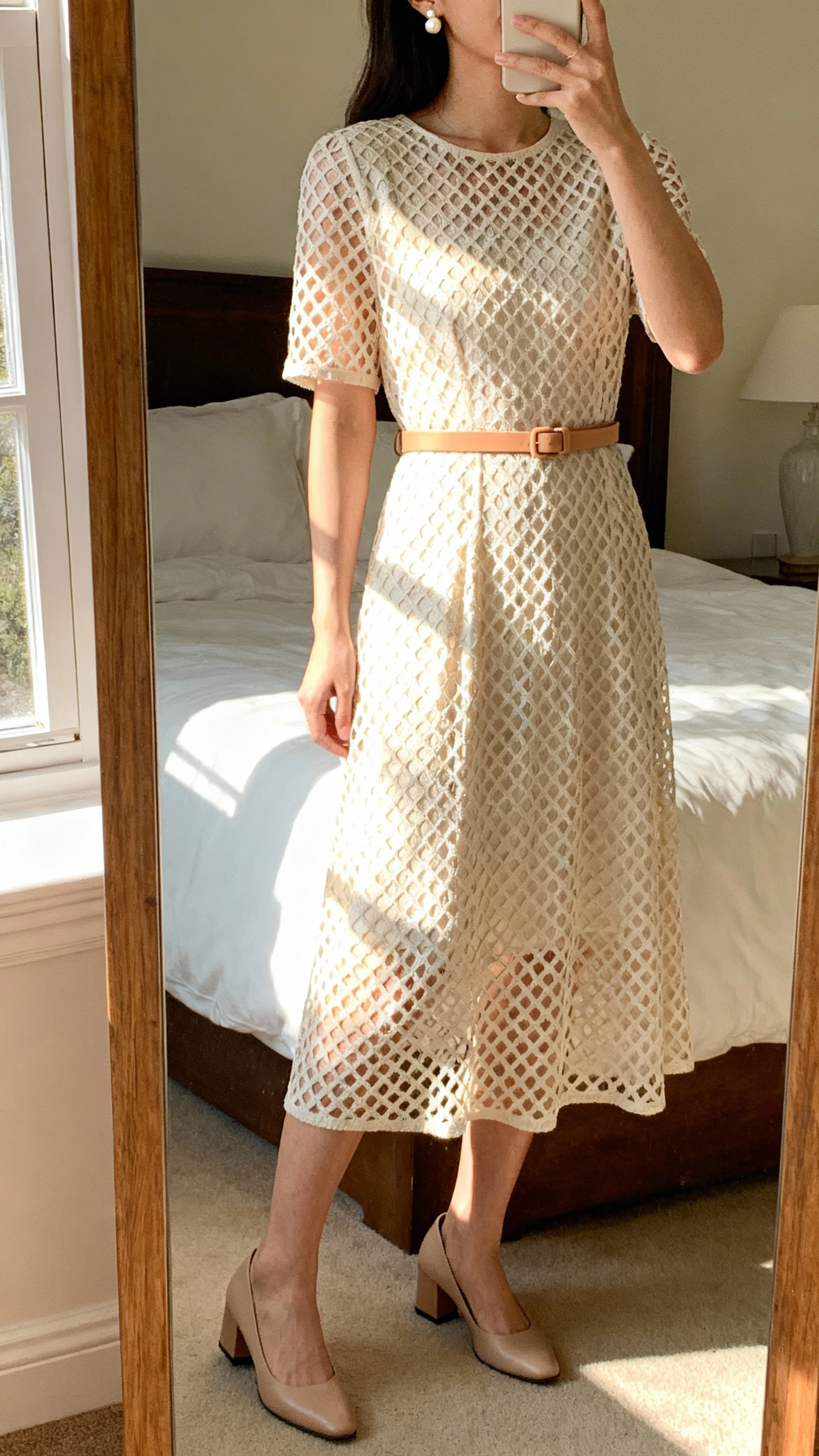 iPhone mirror selfie of a woman wearing a gentle lattice-pattern midi dress that glows in morning light, paired with a thin tan belt, dainty pearl stud earrings, and low block-heel pumps, face not visible, cropped at neck, bedroom mirror, soft morning window light, iPhone photo quality.