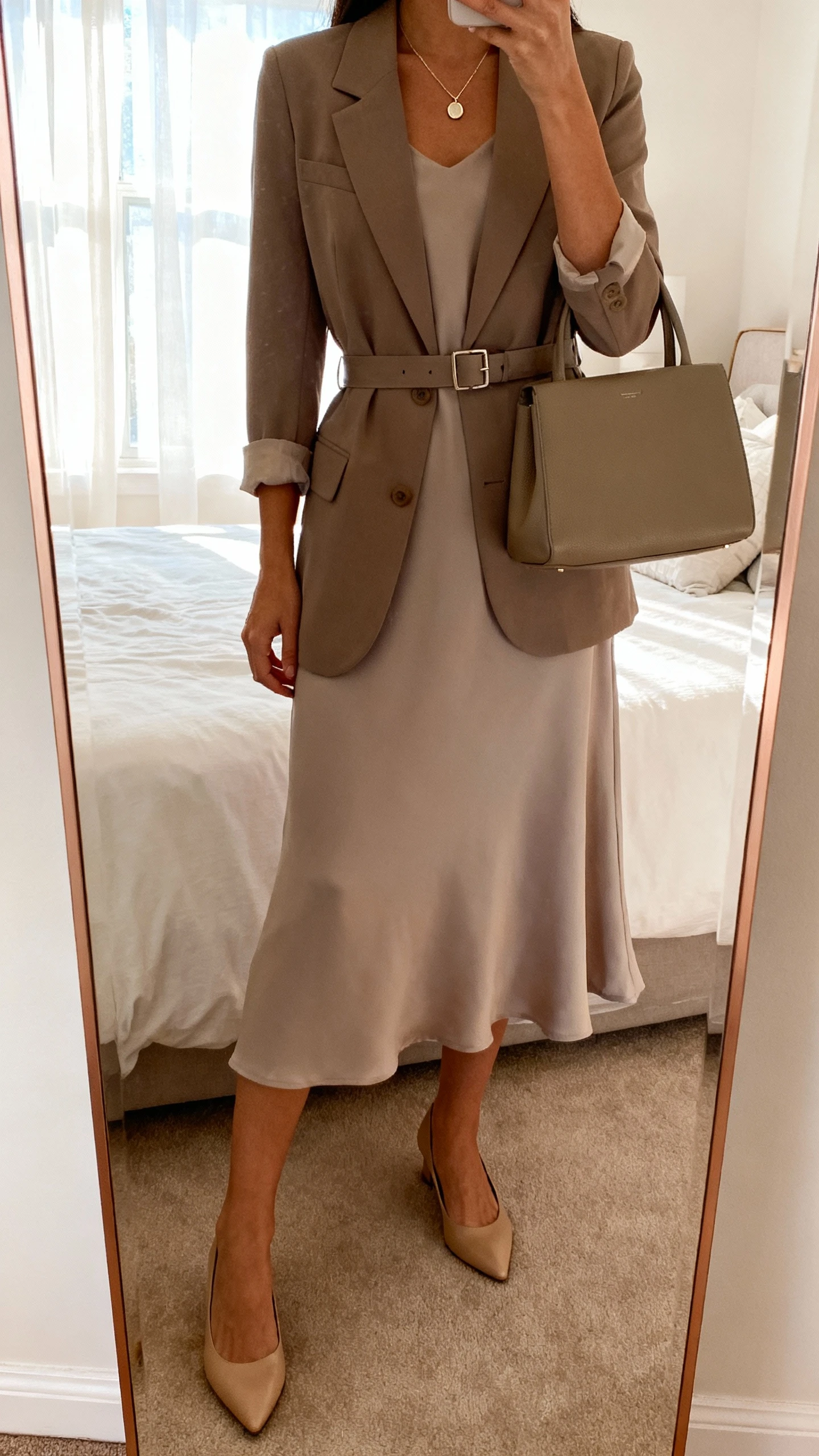 iPhone mirror selfie of a woman wearing a graceful midi dress with a belted blazer, simple pumps, delicate pendant necklace, and a structured tote, face not visible, cropped at neck, bedroom mirror, soft morning window light, iPhone photo quality.