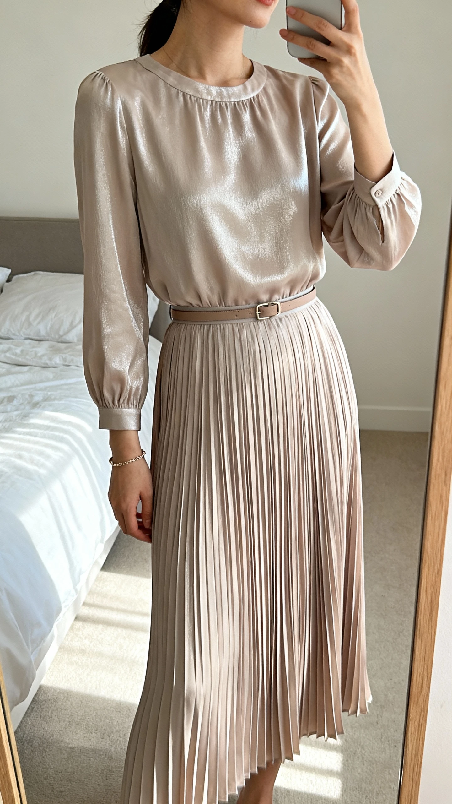 iPhone mirror selfie of a woman wearing a graceful pleated skirt dress with a modest bodice, long sleeves, and a subtle sheen, styled with a slim belt and delicate bracelet, face not visible, cropped at shoulders, bedroom mirror, soft daylight, iPhone photo quality