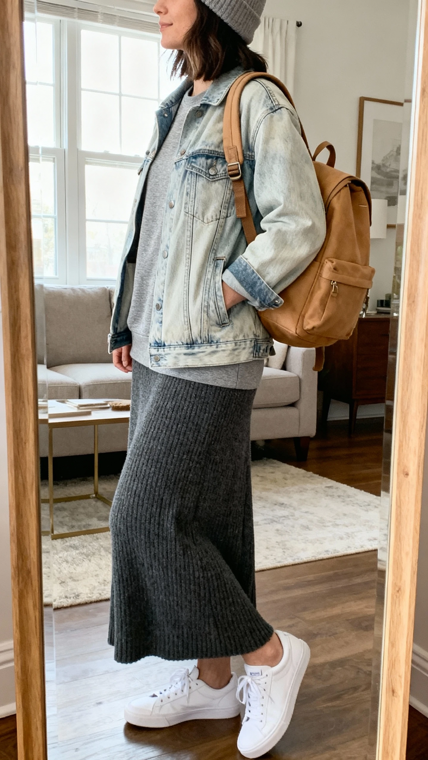 iPhone mirror selfie of a woman wearing a heather gray crewneck under a light-wash denim jacket with a soft charcoal knit maxi skirt, white sneakers, tan backpack, and beanie, face not visible, side angle, living room full-length mirror, natural window light, iPhone photo quality.