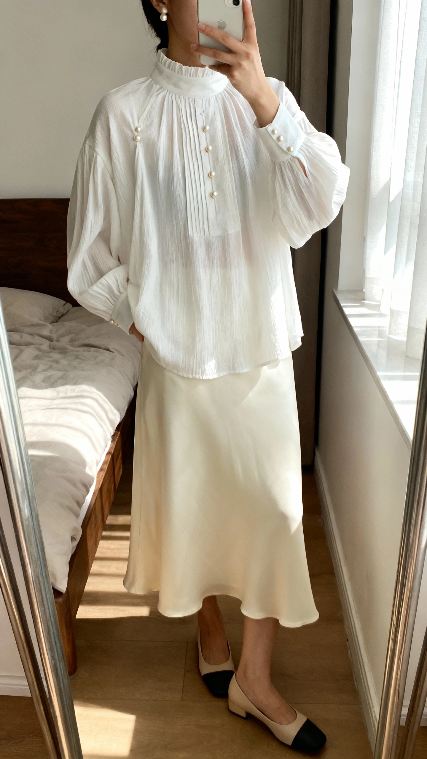iPhone mirror selfie of a woman wearing a high-neck loose-fit white blouse with soft pleats, subtle pearl-button accents, and a flowing midi skirt in cream with simple low block heels and a small pearl-stud earring detail, face not visible, cropped at neck, bedroom mirror, morning window light, iPhone photo quality.