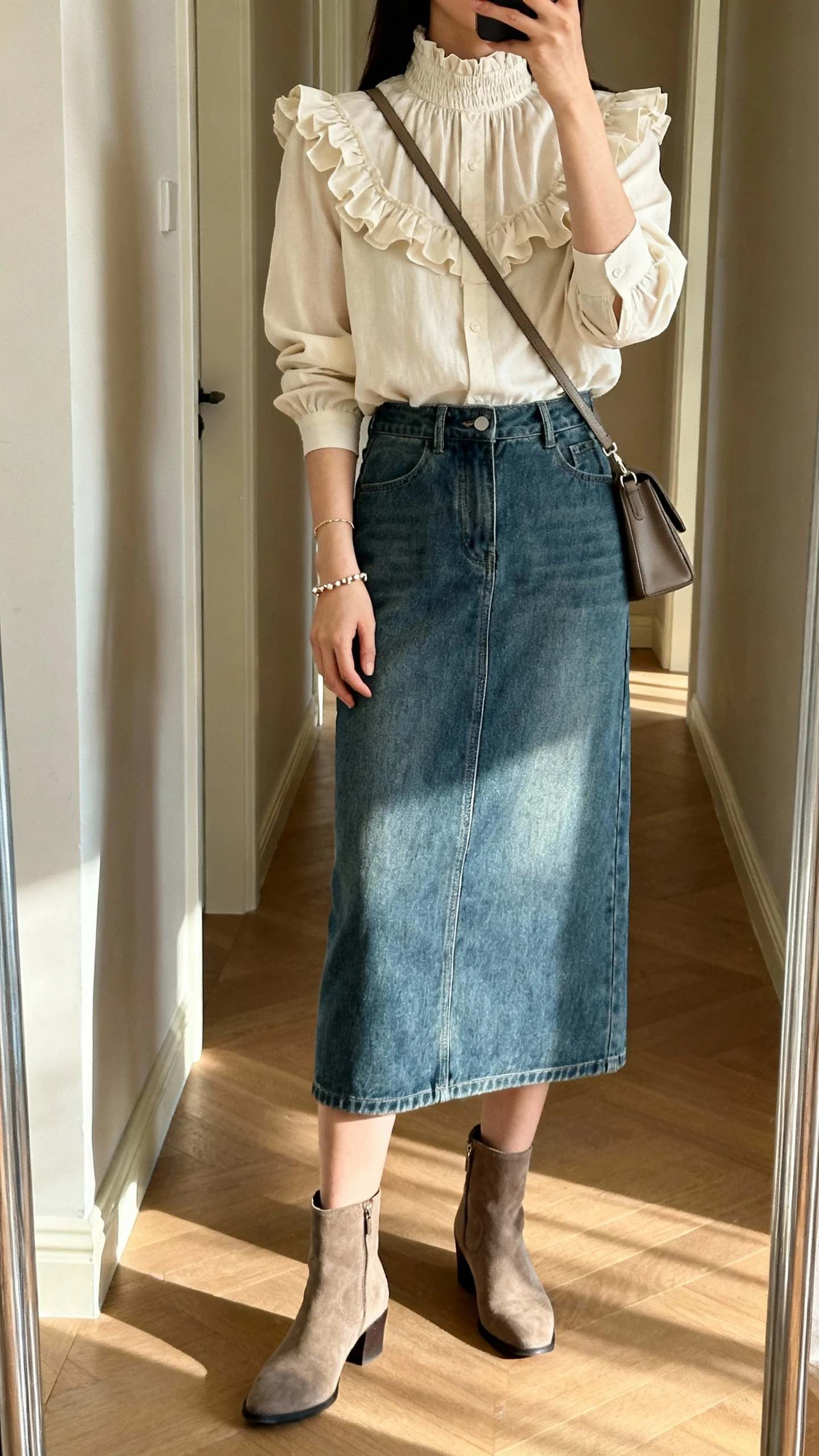 iPhone mirror selfie of a woman wearing a high-neck ruffled blouse paired with a denim midi skirt, ankle boots, dainty bracelet, and a crossbody bag; face not visible, cropped at neck, full-length mirror in hallway, afternoon natural light, iPhone photo quality.