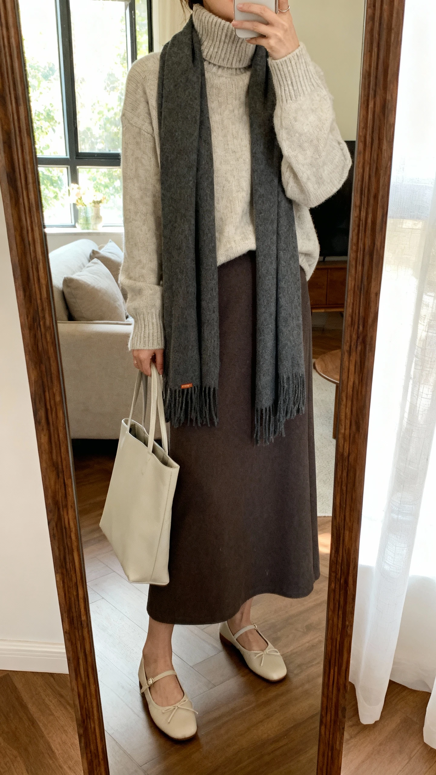 iPhone mirror selfie of a woman wearing a high-neck knit sweater layered over a maxi skirt, cozy scarf, ballet flats, and a simple tote; face not visible, cropped at shoulders, living-room mirror, soft window light, iPhone photo quality.