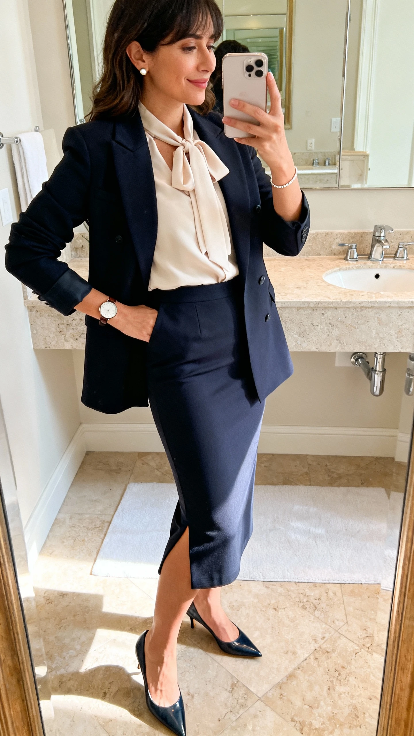 iPhone mirror selfie of a woman wearing a high-waisted midi pencil skirt (back slit only), a soft tie-neck blouse, a structured blazer, pointed-toe kitten heels, a simple watch, stud earrings, and a delicate bracelet, face not visible, bathroom mirror, indoor morning light, iPhone photo quality.