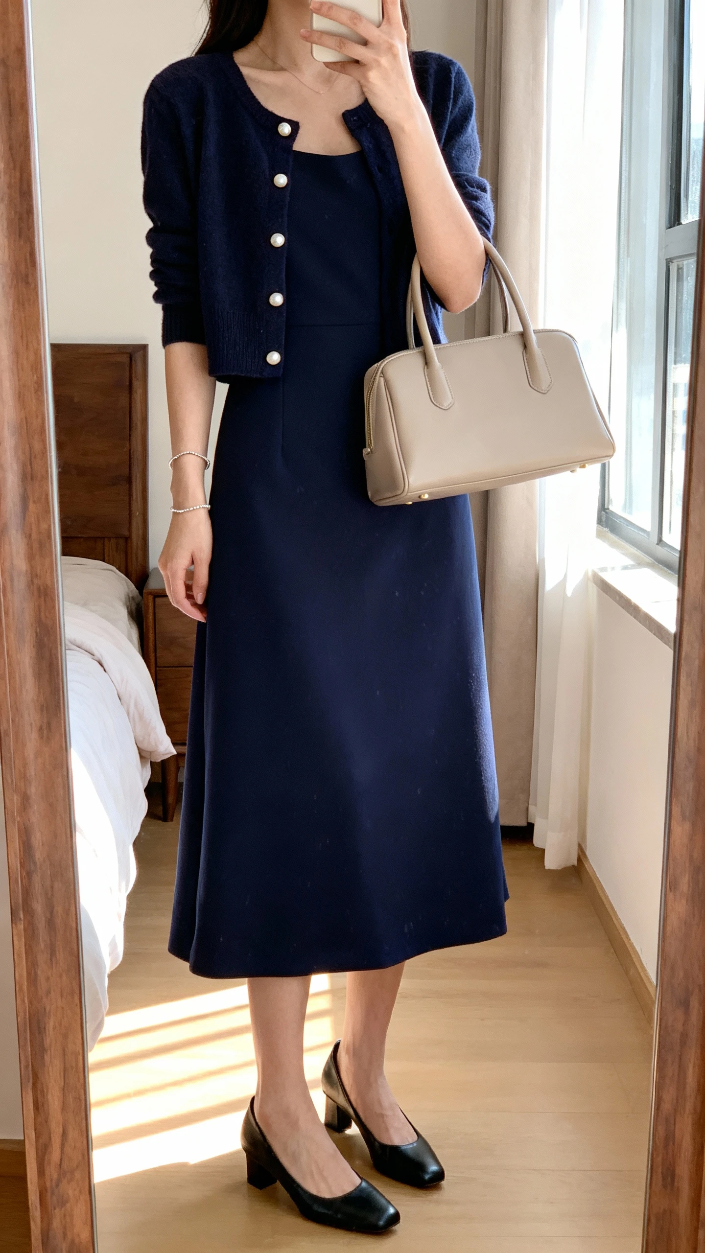 iPhone mirror selfie of a woman wearing a knee-length A-line dress in navy with a cropped cardigan, classic black pumps, pearl studs, a delicate bracelet, and a structured neutral top-handle bag, face not visible, cropped at neck, bedroom mirror with morning window light, iPhone photo quality.
