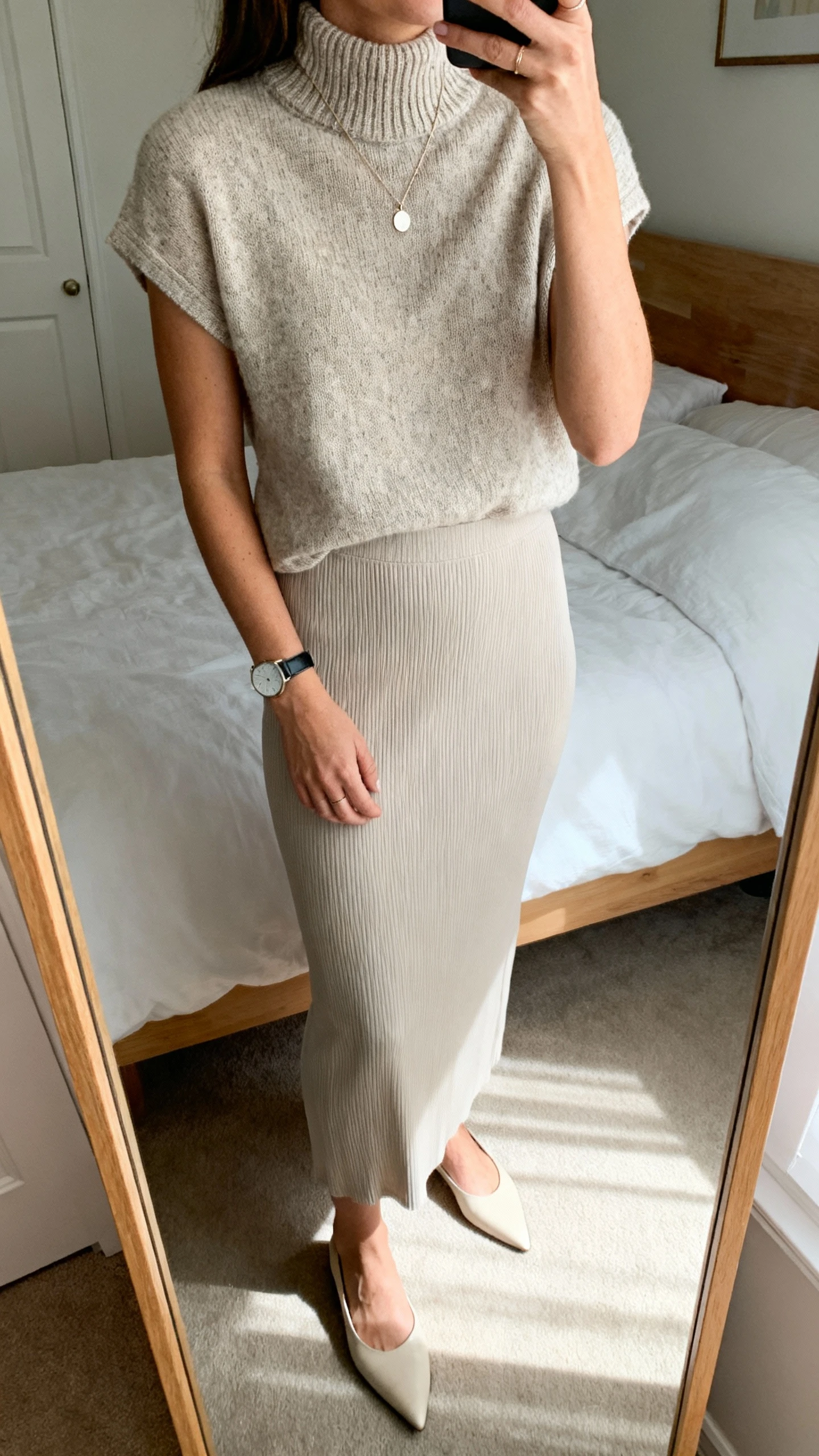 iPhone mirror selfie of a woman wearing a knit mock-neck top with a column skirt, minimal pendant necklace, sleek watch, and pointed flats, face not visible, cropped at shoulders, bedroom mirror, gentle afternoon light, iPhone photo quality.