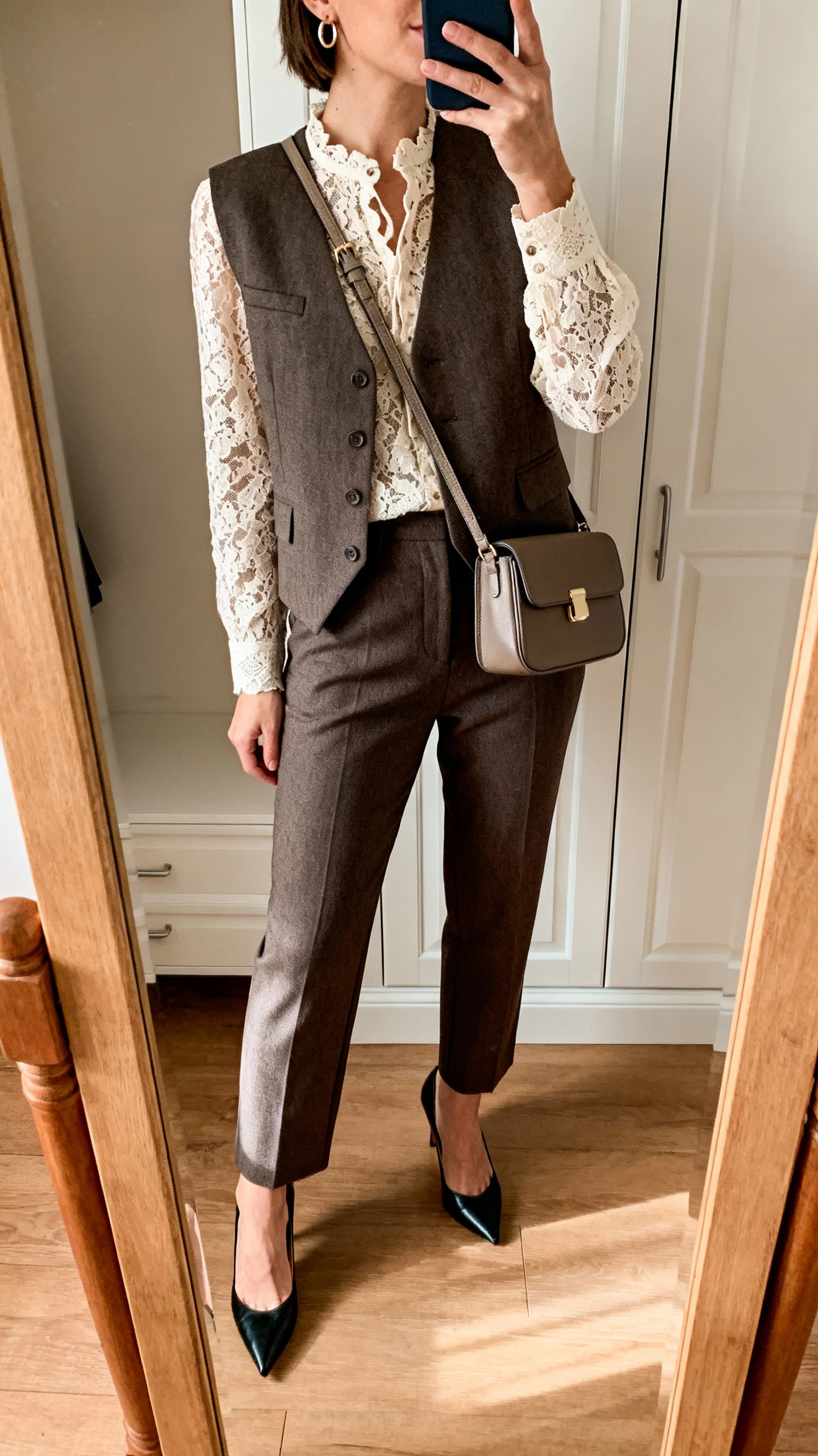 iPhone mirror selfie of a woman wearing a lace-detail blouse layered under a tailored vest with straight-leg trousers, kitten heels, a structured mini shoulder bag, and small hoop earrings, face not visible, cropped at neck, closet mirror, warm indoor lighting, iPhone photo quality.