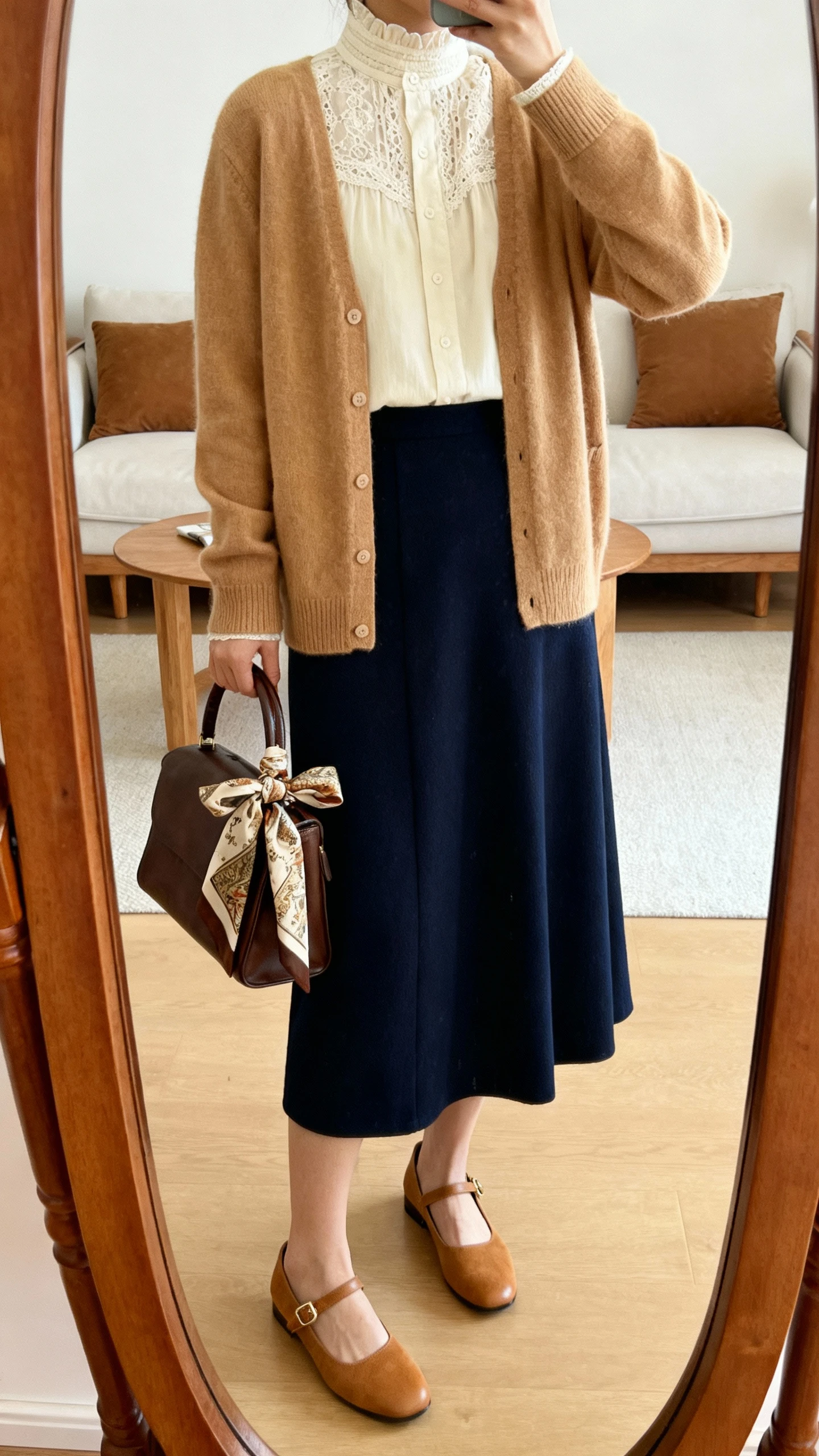 iPhone mirror selfie of a woman wearing a lace-trim high-neck blouse in cream under a classic camel cardigan, paired with a navy midi skirt, tan Mary Janes, and a vintage-style scarf tied at the handle of a satchel, face not visible, cropped at shoulders, living room full-length mirror, warm indoor lighting, iPhone photo quality.