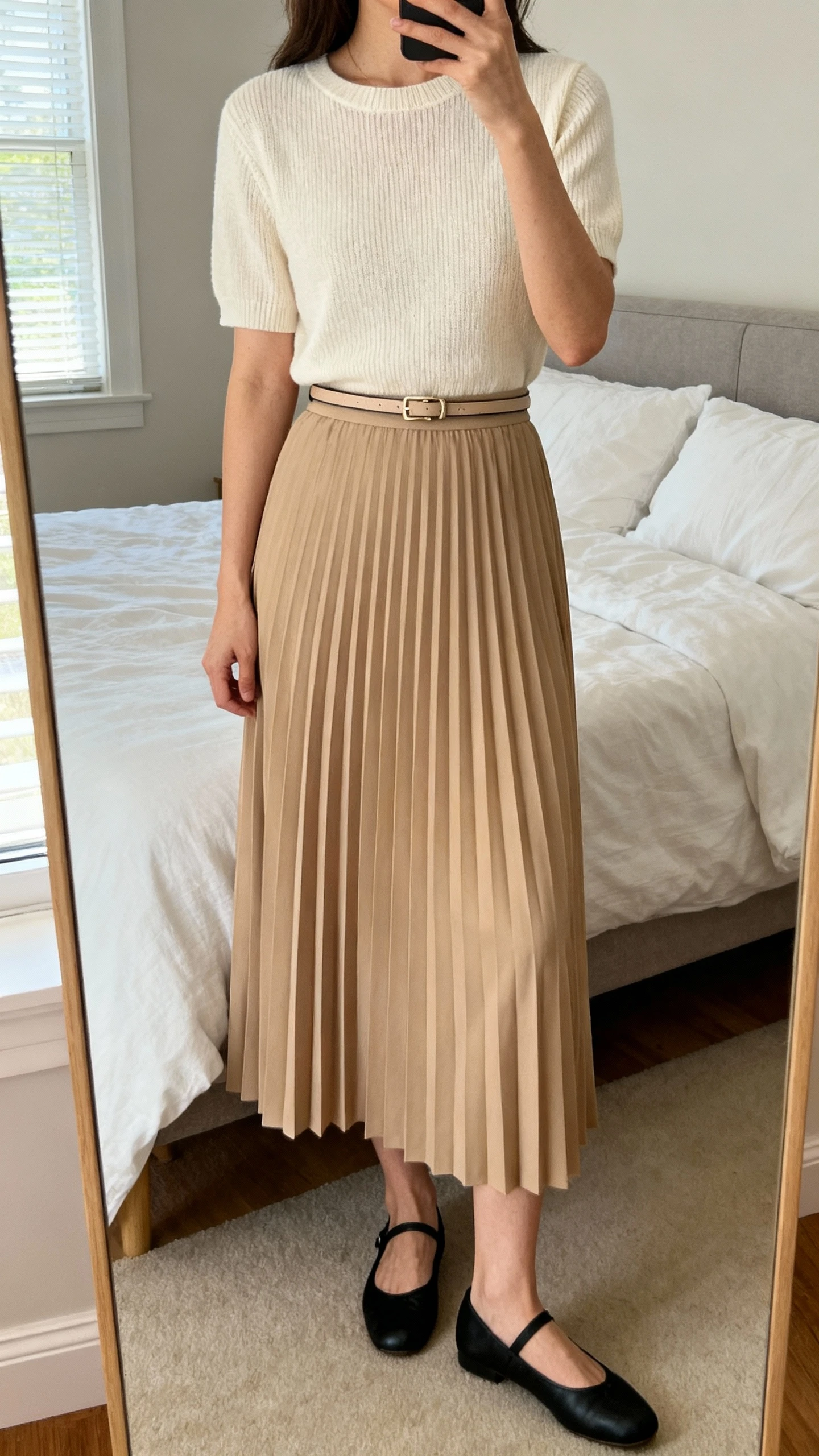 iPhone mirror selfie of a woman wearing a latte pleated midi skirt with a cream short-sleeve fine-knit top, black ballet flats, and a slim belt defining the waist, face not visible, cropped at neck, bedroom mirror, natural window light, iPhone photo quality.