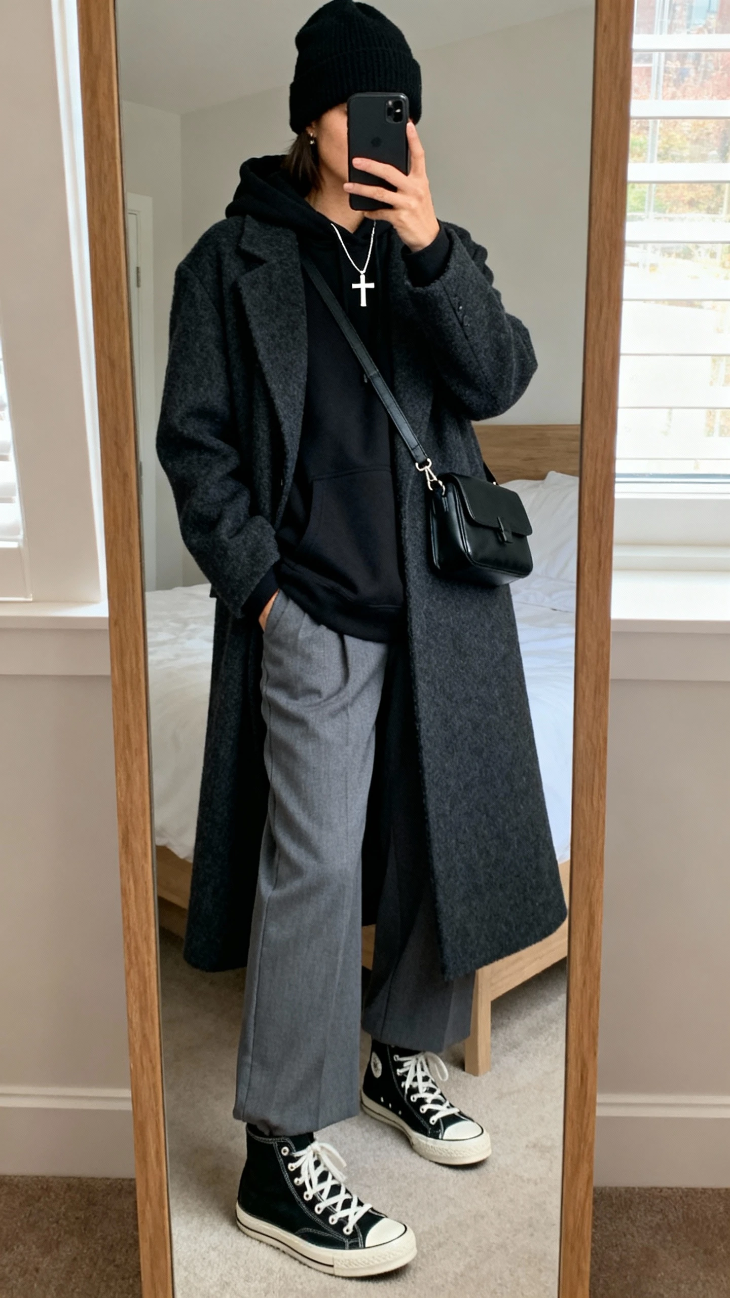 iPhone mirror selfie of a woman wearing a layered monochrome set: black oversized hoodie under a charcoal longline wool coat, gray wide-leg trousers, black high-top sneakers, black beanie, minimal silver cross necklace, small black crossbody bag; face not visible, cropped at neck, bedroom mirror, natural window light, iPhone photo quality.