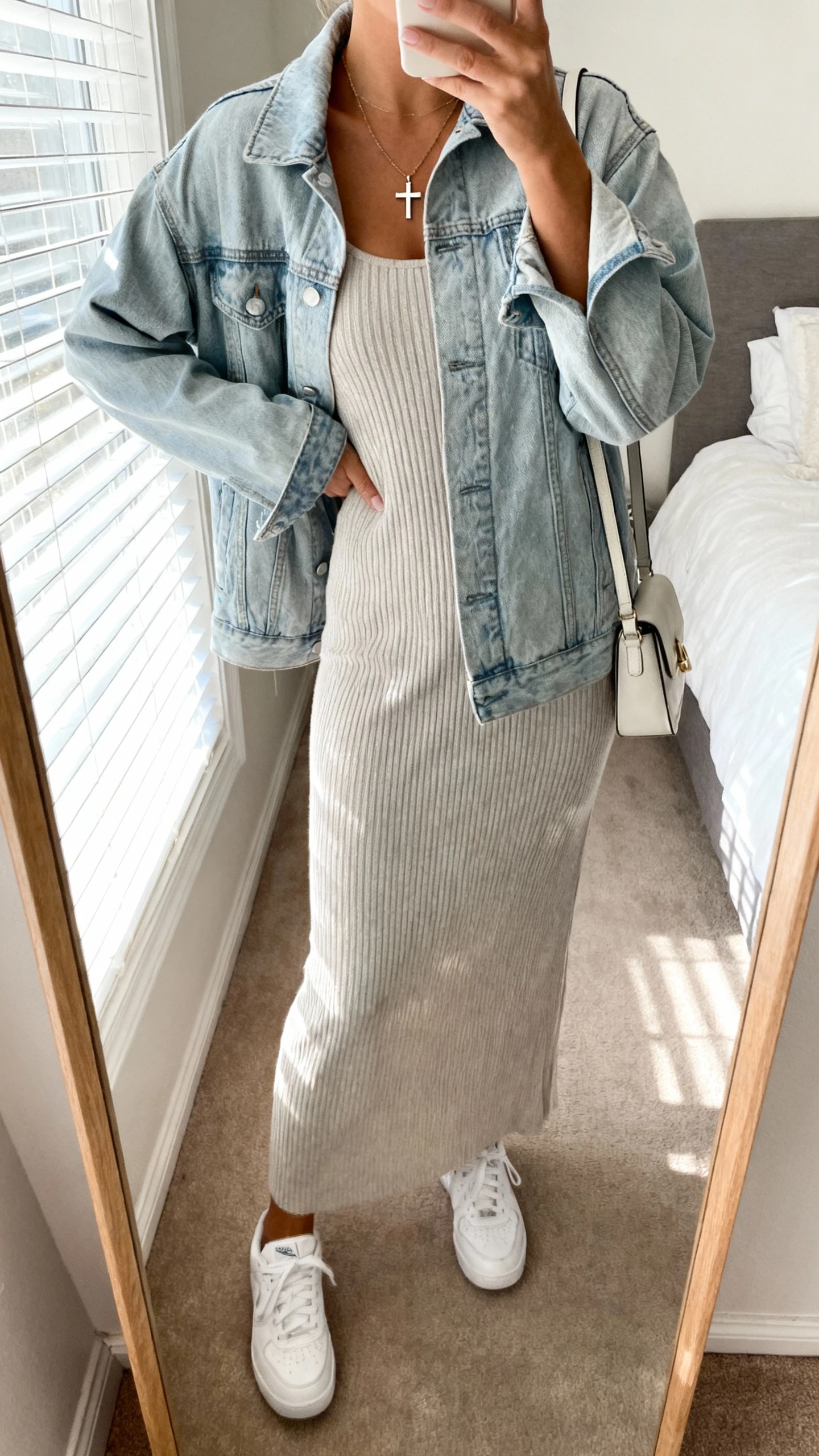 iPhone mirror selfie of a woman wearing a light denim jacket over a ribbed knit maxi dress with clean white sneakers, simple cross pendant necklace, small shoulder bag, face not visible, cropped at neck, bedroom mirror, soft morning window light, iPhone photo quality.