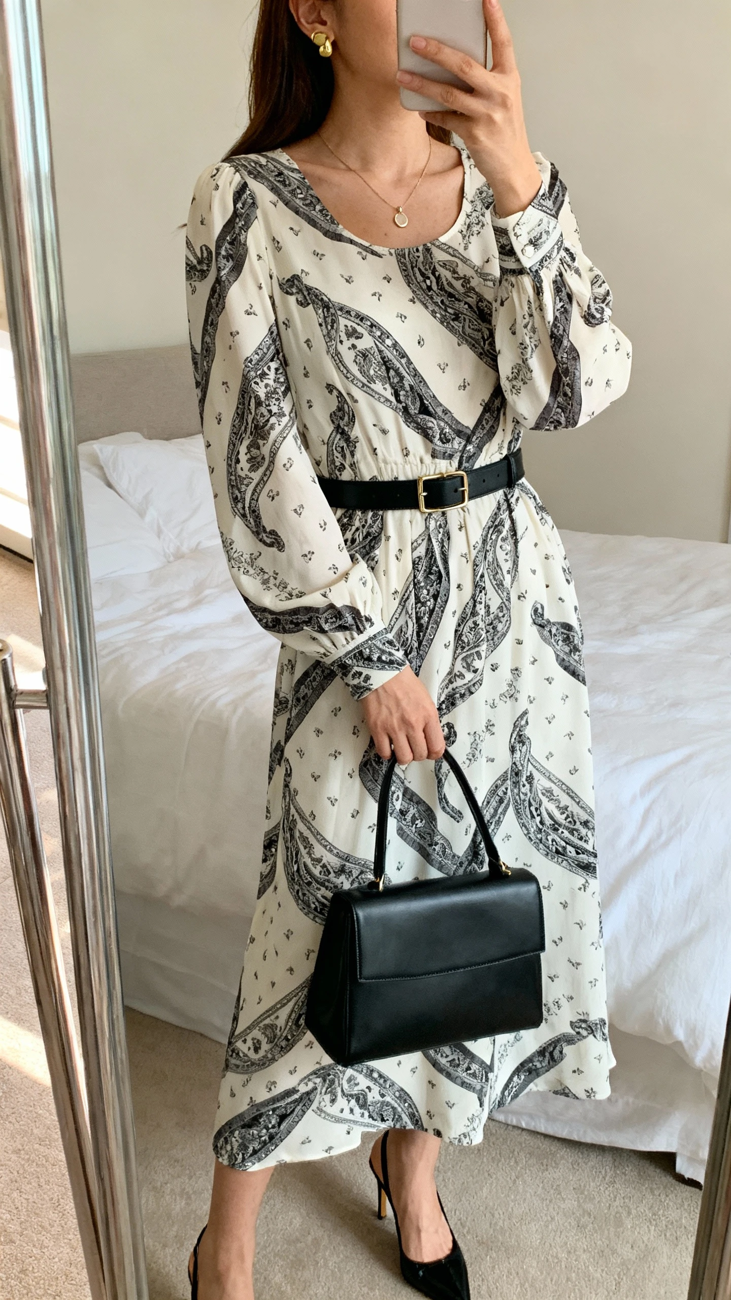 iPhone mirror selfie of a woman wearing a long-sleeve scarf-print midi dress with a modest neckline, a slim black leather belt, kitten-heel slingbacks, a structured black satchel, gold stud earrings, and a dainty pendant necklace, face not visible, cropped at shoulders, bedroom mirror, soft natural light, iPhone photo quality.