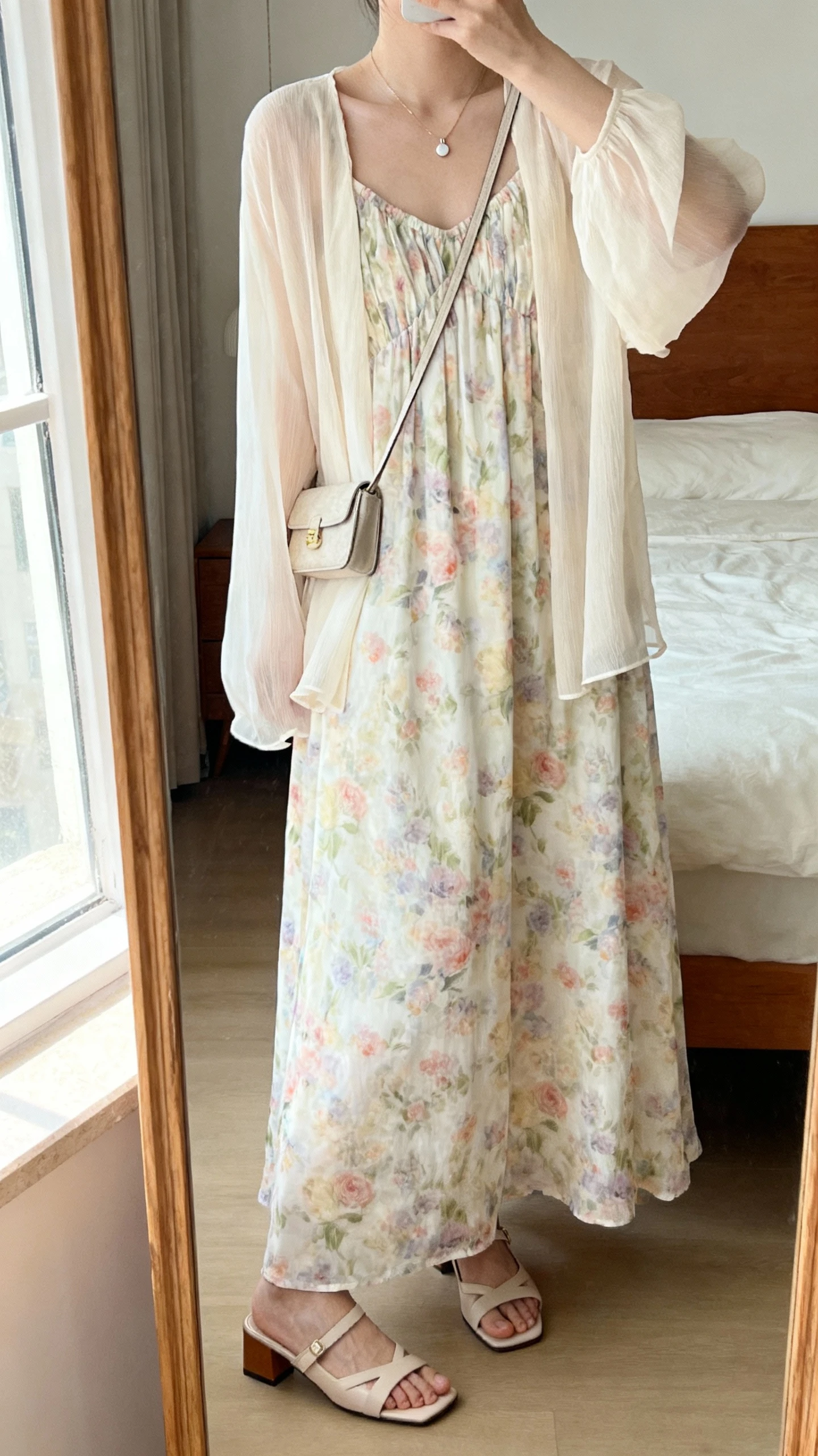 iPhone mirror selfie of a woman wearing a loose-fit floral maxi dress with soft pastel blooms, light chiffon cardigan layered over, delicate pendant necklace, small crossbody bag, and low block-heel sandals, face not visible, cropped at neck, bedroom mirror, morning window light, iPhone photo quality