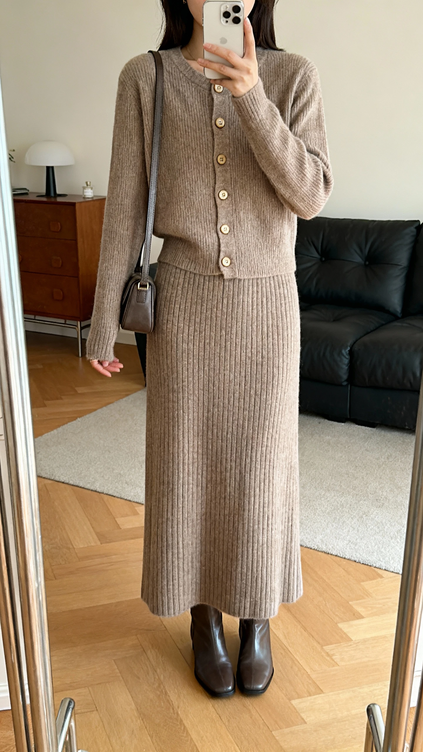 iPhone mirror selfie of a woman wearing a matching knit set: long ribbed skirt and buttoned cardigan, with sleek ankle boots and a small shoulder bag, face not visible, cropped at shoulders, living room mirror, warm indoor lighting, iPhone photo quality.