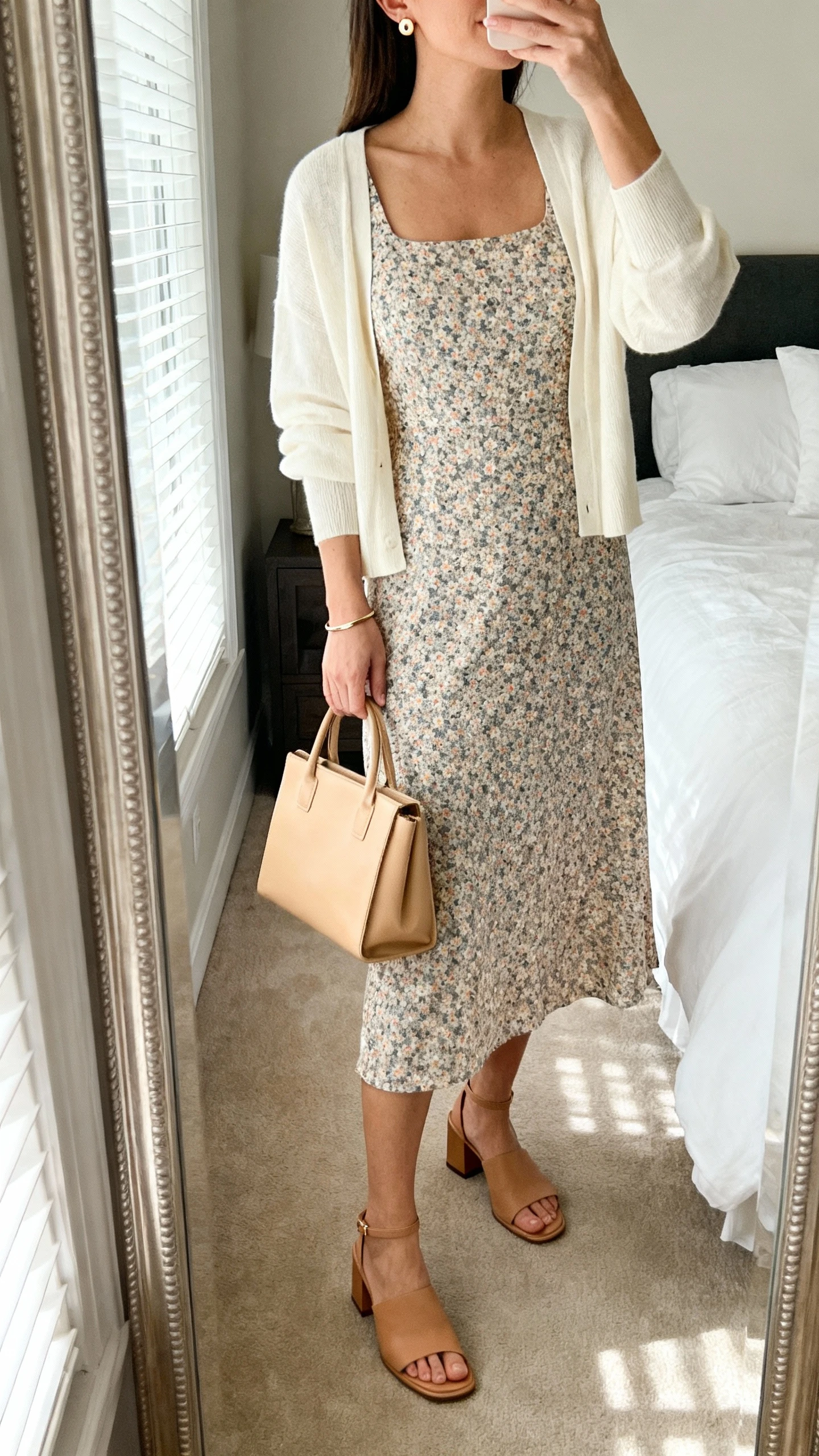 iPhone mirror selfie of a woman wearing a micro-floral midi dress with a modest scoop/square neckline, a lightweight cream cardigan, low camel block-heel sandals or closed-toe flats, simple gold stud earrings, a thin bracelet, and a structured sand tote; face not visible, cropped at neck, bedroom mirror, soft morning window light, iPhone photo quality.