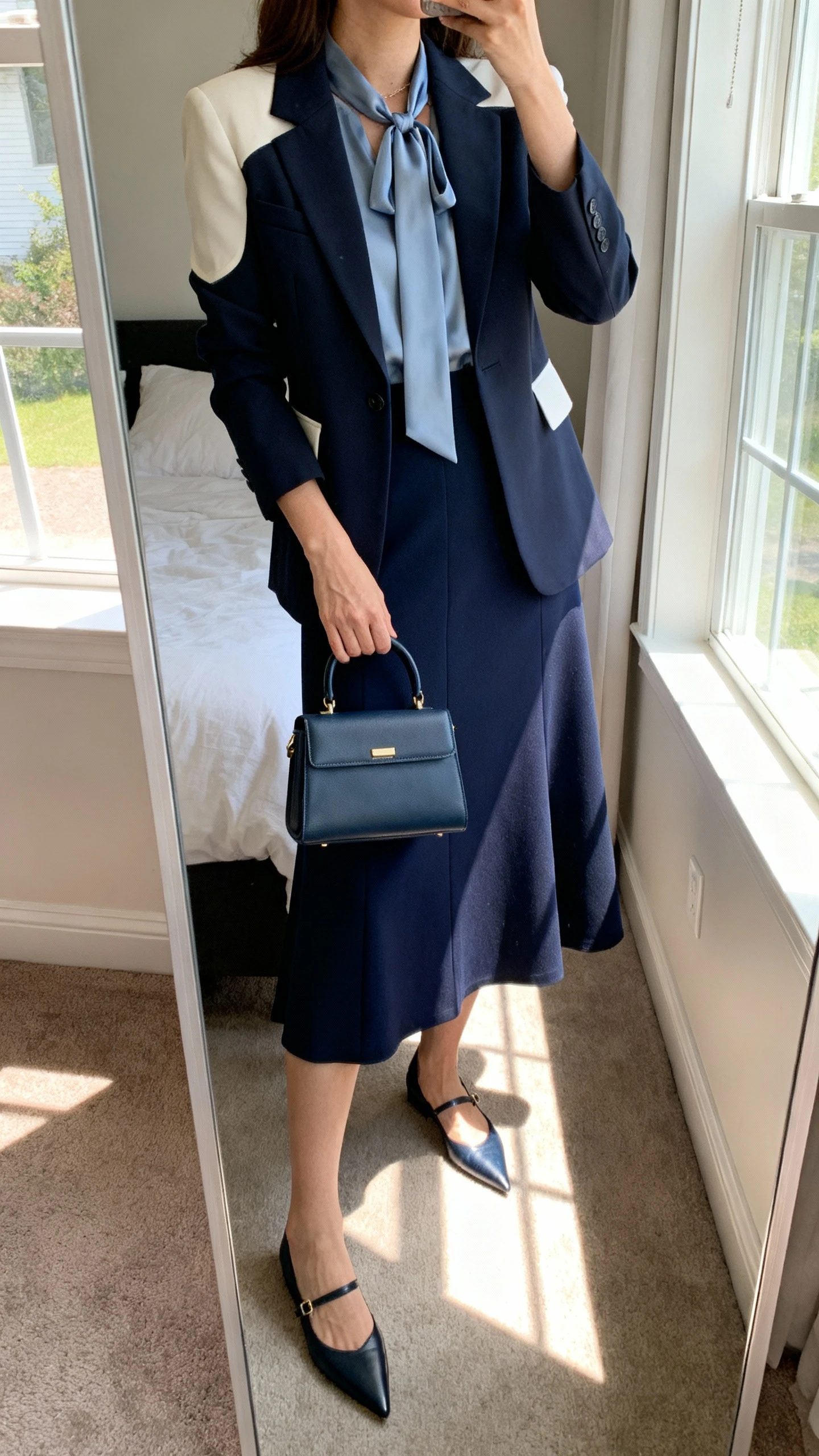 iPhone mirror selfie of a woman wearing a mid-length A-line skirt in navy, a single-breasted tailored blazer with light shoulder structure, a silk tie-neck blouse with a modest neckline, pointed-toe flats, and a structured top-handle bag, face not visible, cropped at neck, bedroom mirror, natural window light, iPhone photo quality.