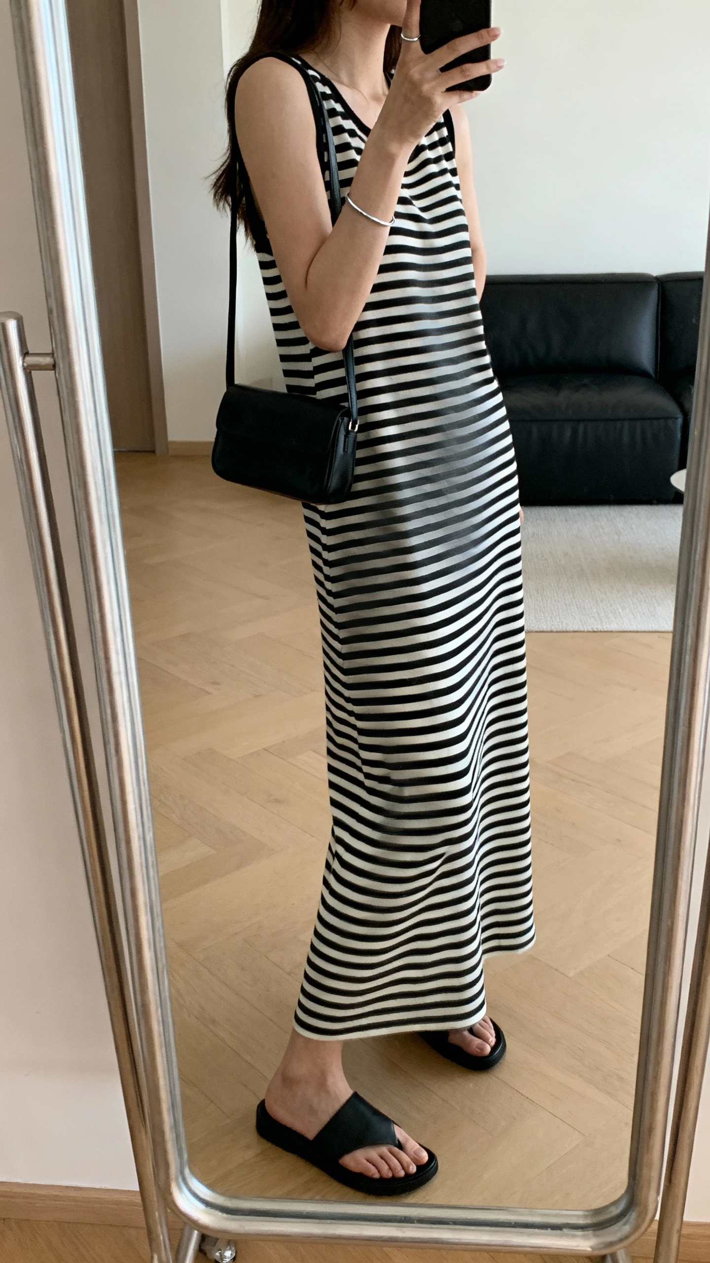 iPhone mirror selfie of a woman wearing a minimalist monochrome black-and-white striped maxi dress, sleek black slide sandals, small black shoulder bag, thin silver bracelet, face not visible, side angle, living room full-length mirror, natural daylight, iPhone photo quality.