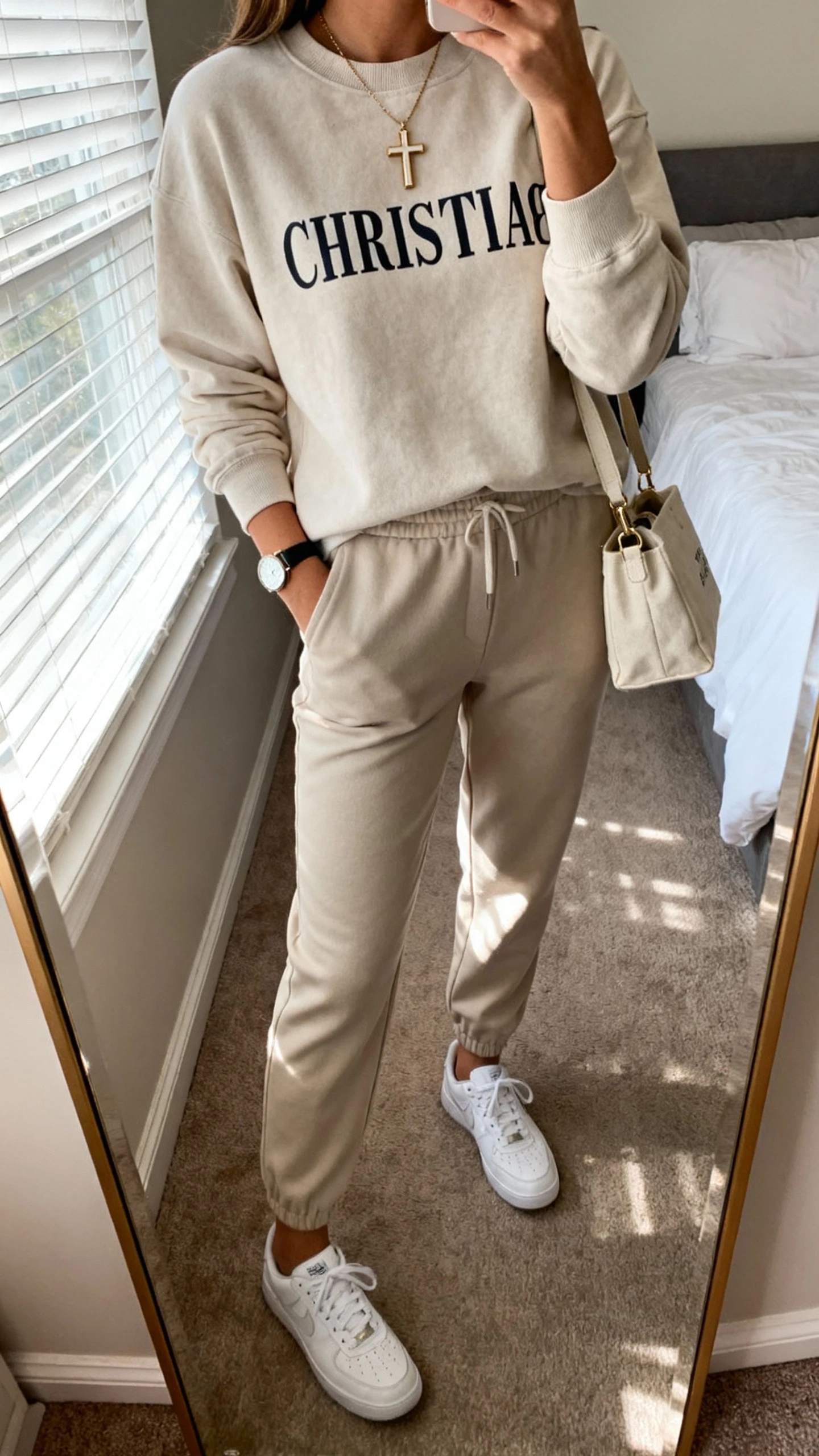 iPhone mirror selfie of a woman wearing a modest Christian crewneck with tailored joggers, clean white sneakers, cross pendant necklace, small tote bag, and a simple watch; face not visible, cropped at neck, bedroom mirror, morning window light, iPhone photo quality.