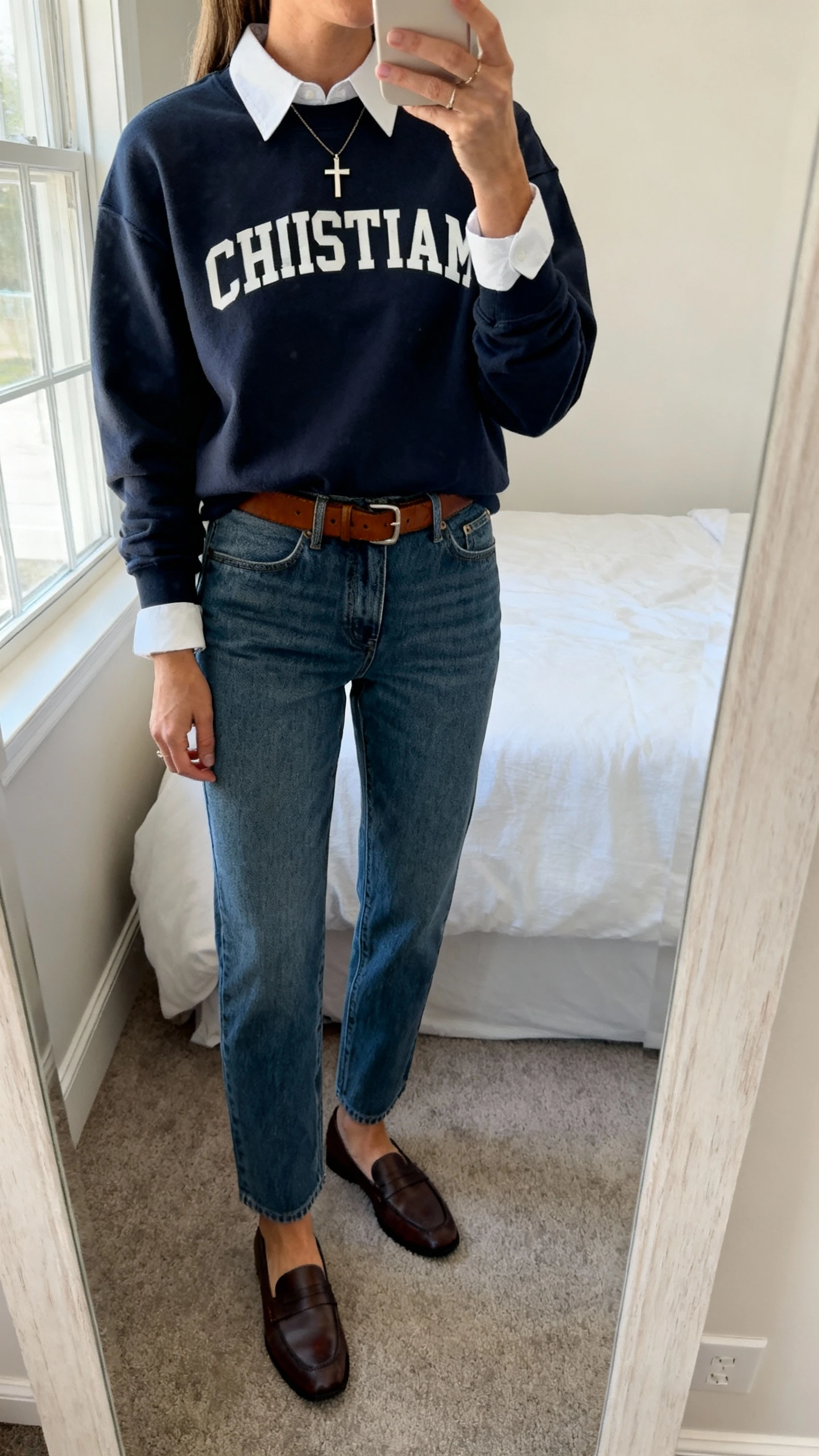 iPhone mirror selfie of a woman wearing a modest Christian crewneck layered over a white collared shirt with straight-leg jeans, leather belt, loafers, and a simple cross necklace; face not visible, cropped at shoulders, bedroom mirror, natural window light, iPhone photo quality.