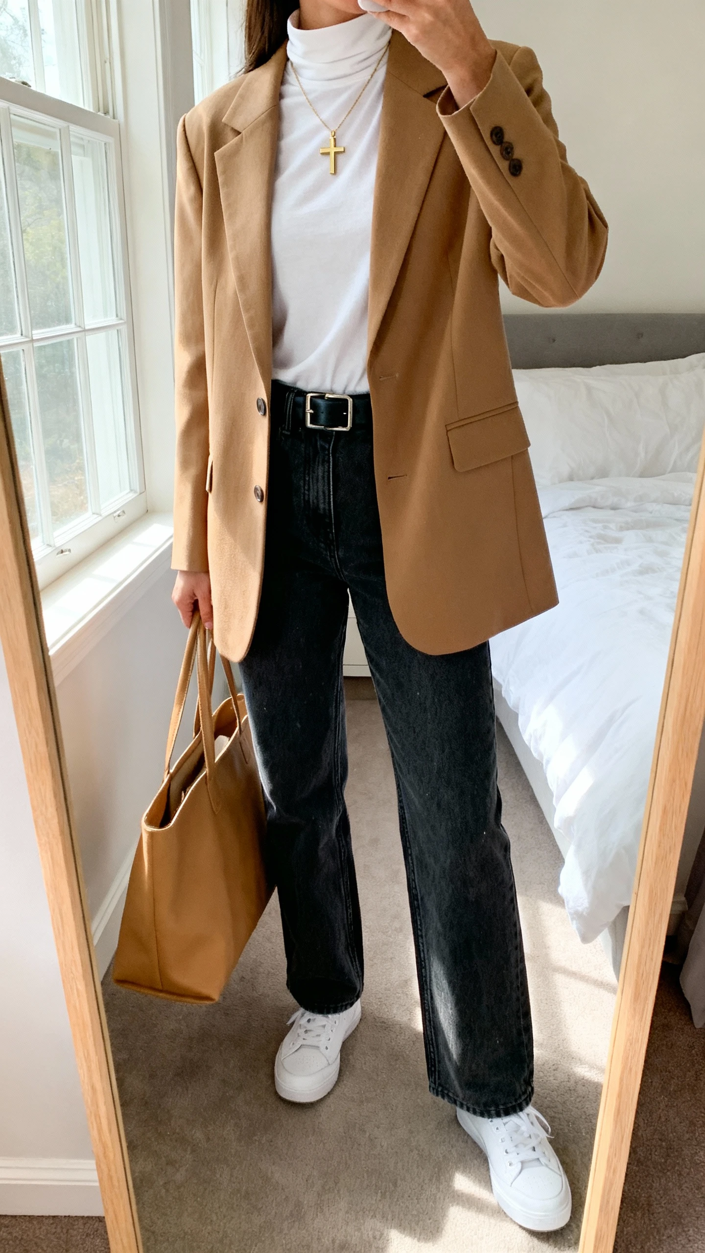 iPhone mirror selfie of a woman wearing a modest dark-wash straight-leg denim, a structured camel blazer over a high-neck white tee, black leather belt, low-top white sneakers, simple gold cross necklace, and a tan tote bag; face not visible, cropped at neck, bedroom mirror, morning window light, iPhone photo quality.