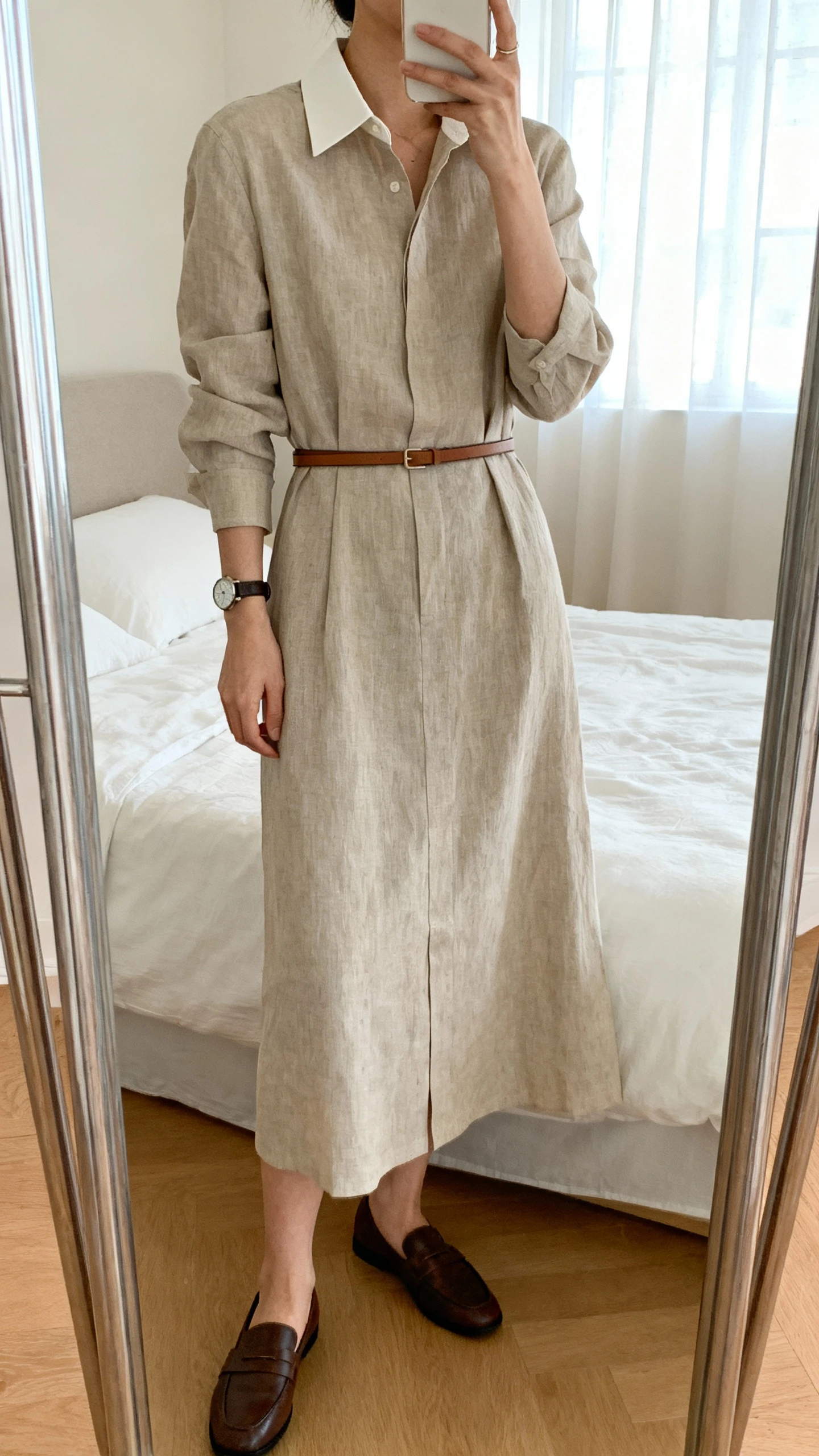 iPhone mirror selfie of a woman wearing a modest tailored linen shirt dress with a crisp collar, thin leather belt, classic watch, and loafers, face not visible, cropped at shoulders, bedroom mirror, natural window light, iPhone photo quality.