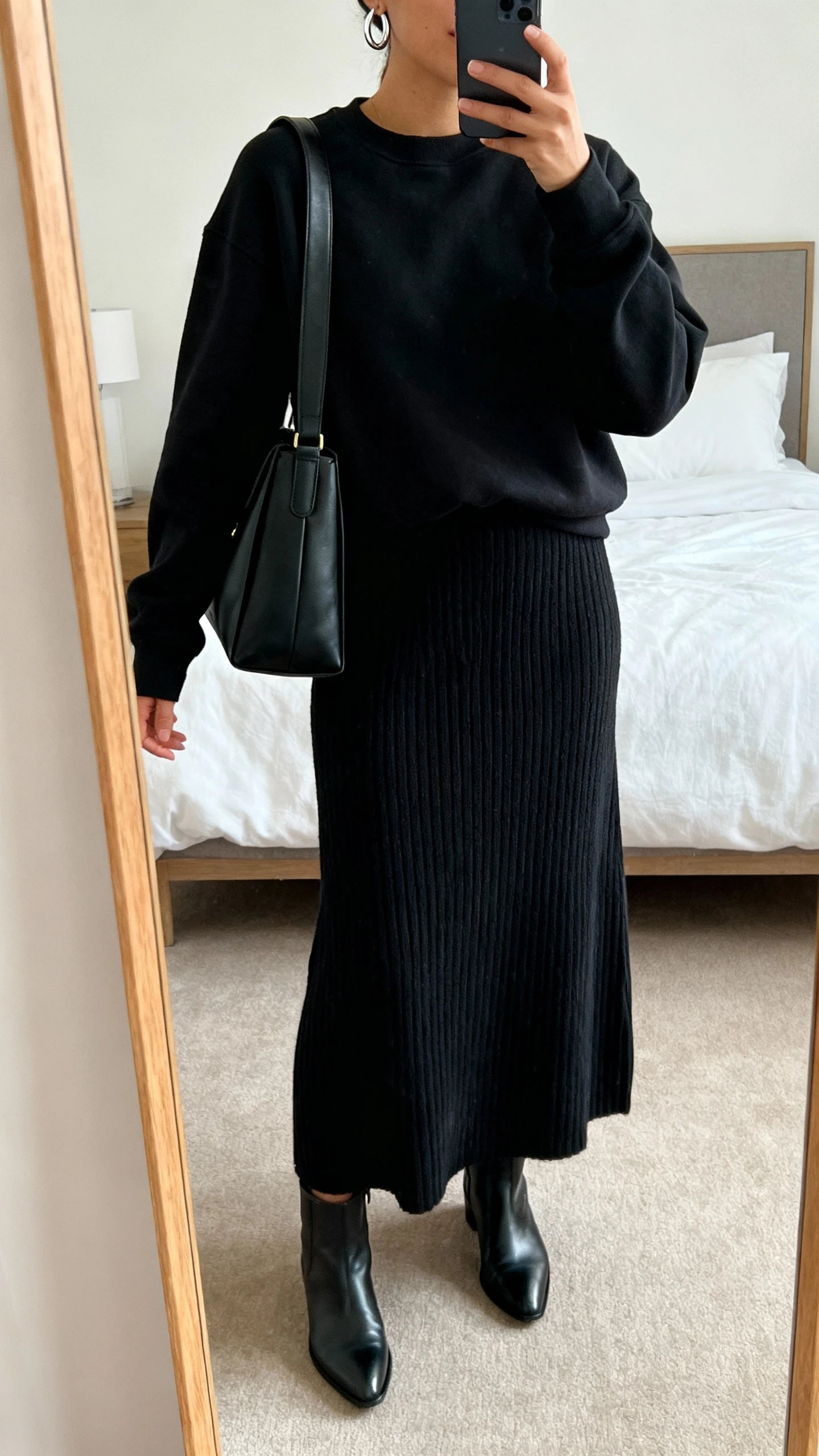 iPhone mirror selfie of a woman wearing a monochrome black crewneck, ribbed knit maxi skirt, structured black shoulder bag, and matte leather ankle boots, subtle silver hoop earrings, face not visible, cropped at shoulders, bedroom mirror, soft natural light, iPhone photo quality.