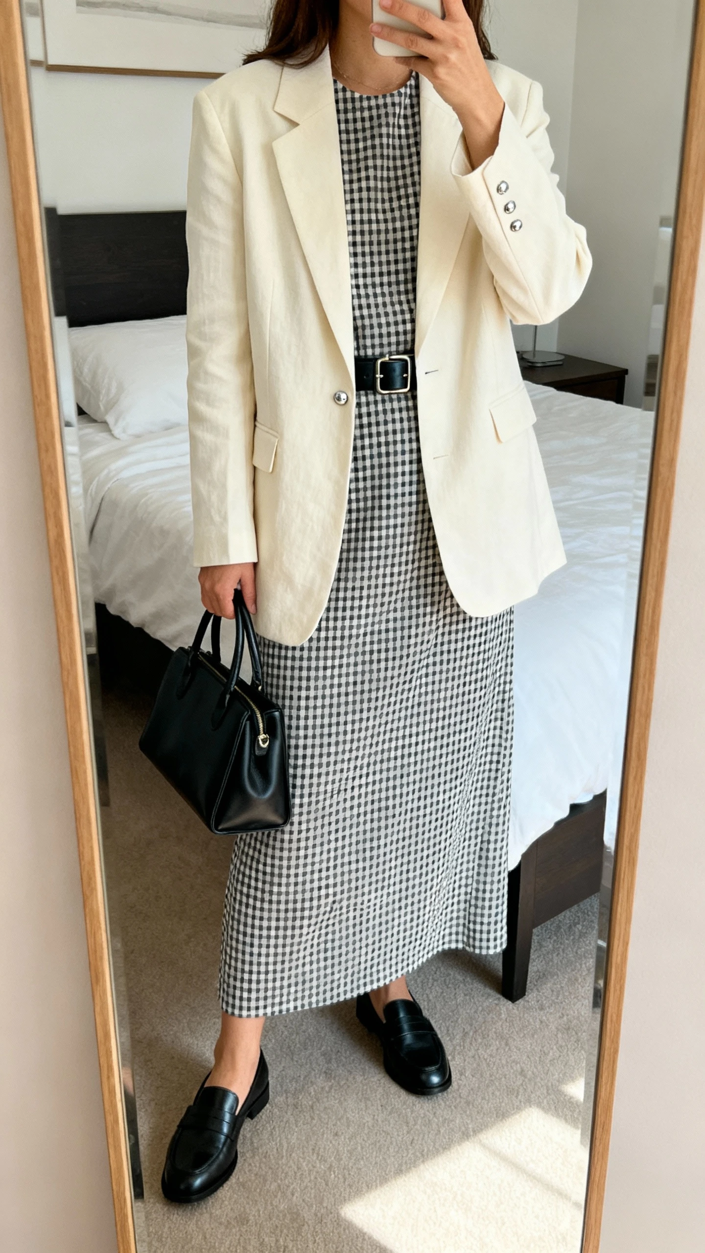 iPhone mirror selfie of a woman wearing a monochrome gingham full-length dress under a soft cream blazer, black leather belt at the waist, black loafers, structured black tote, and minimal silver studs, face not visible, cropped at shoulders, bedroom mirror, natural daylight, iPhone photo quality.