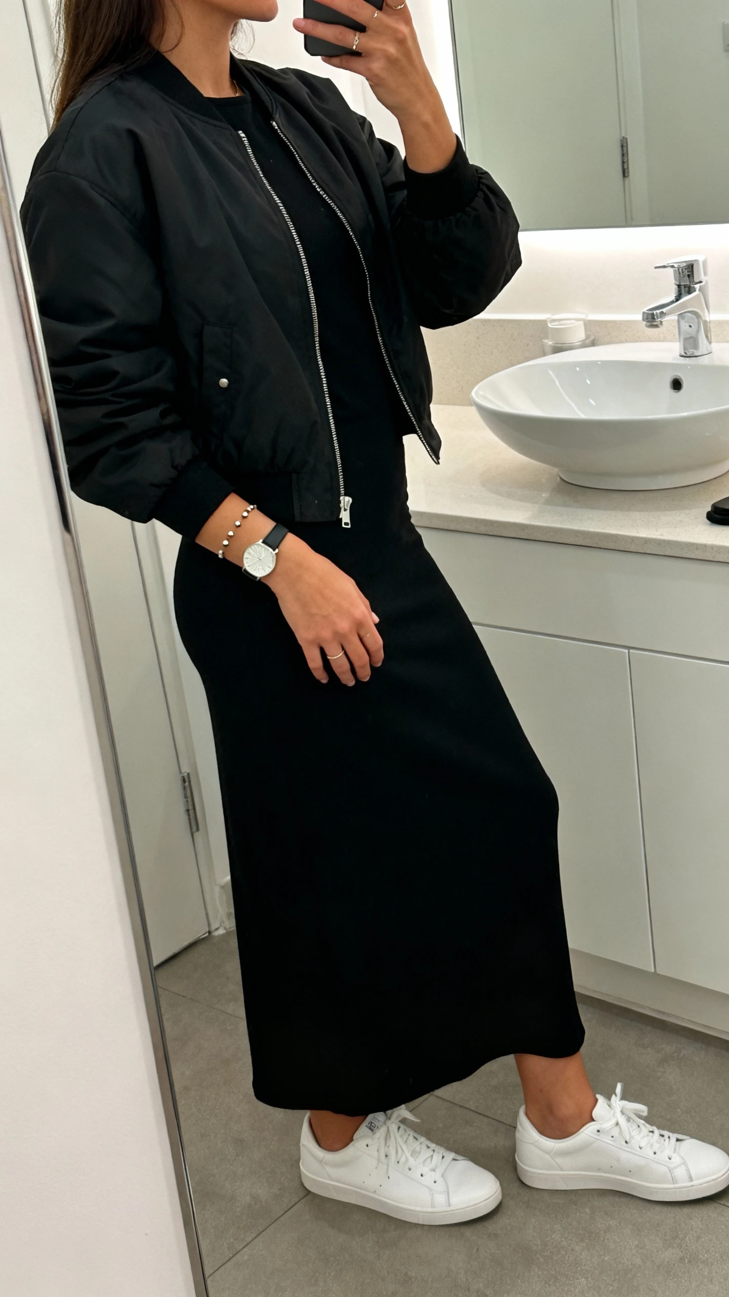 iPhone mirror selfie of a woman wearing a monochrome black maxi dress with a black bomber jacket and crisp white sneakers, simple studs, slim watch, face not visible, side angle, bathroom mirror, indoor lighting, iPhone photo quality.