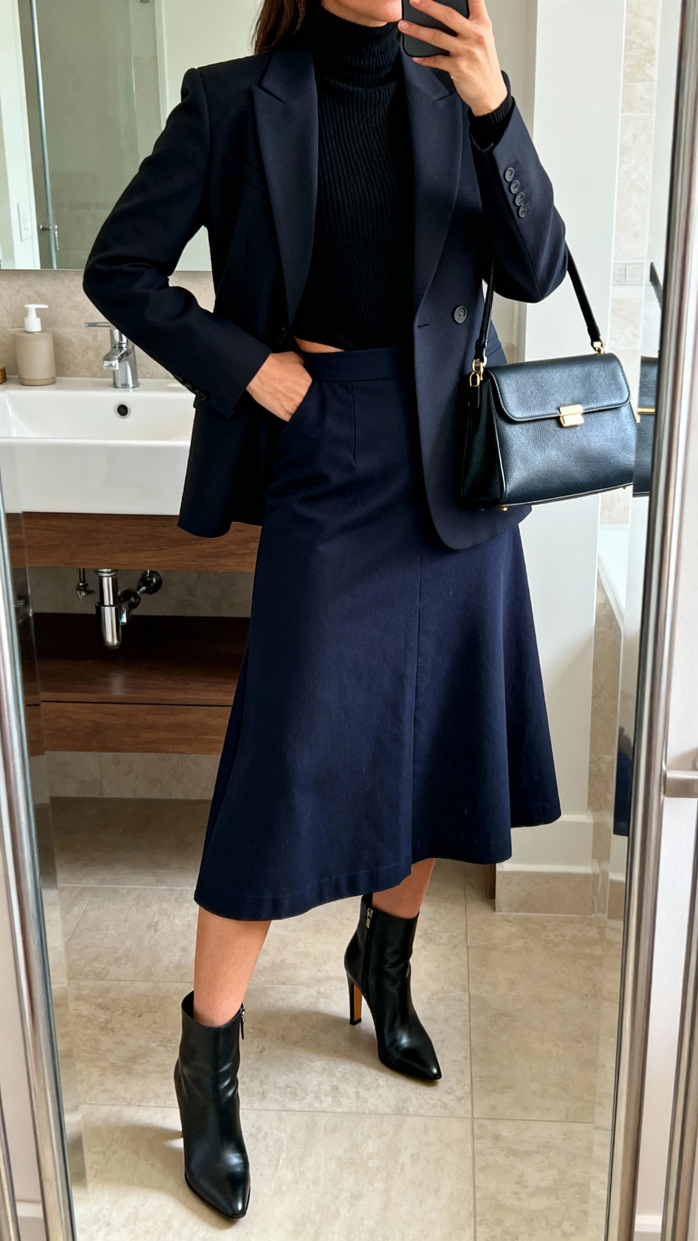 iPhone mirror selfie of a woman wearing a navy A-line midi skirt in twill, a single-breasted tailored blazer nipped at the waist over a fine-gauge black turtleneck, heeled ankle boots, and a structured satchel; face not visible, bathroom mirror, indoor lighting, iPhone photo quality.