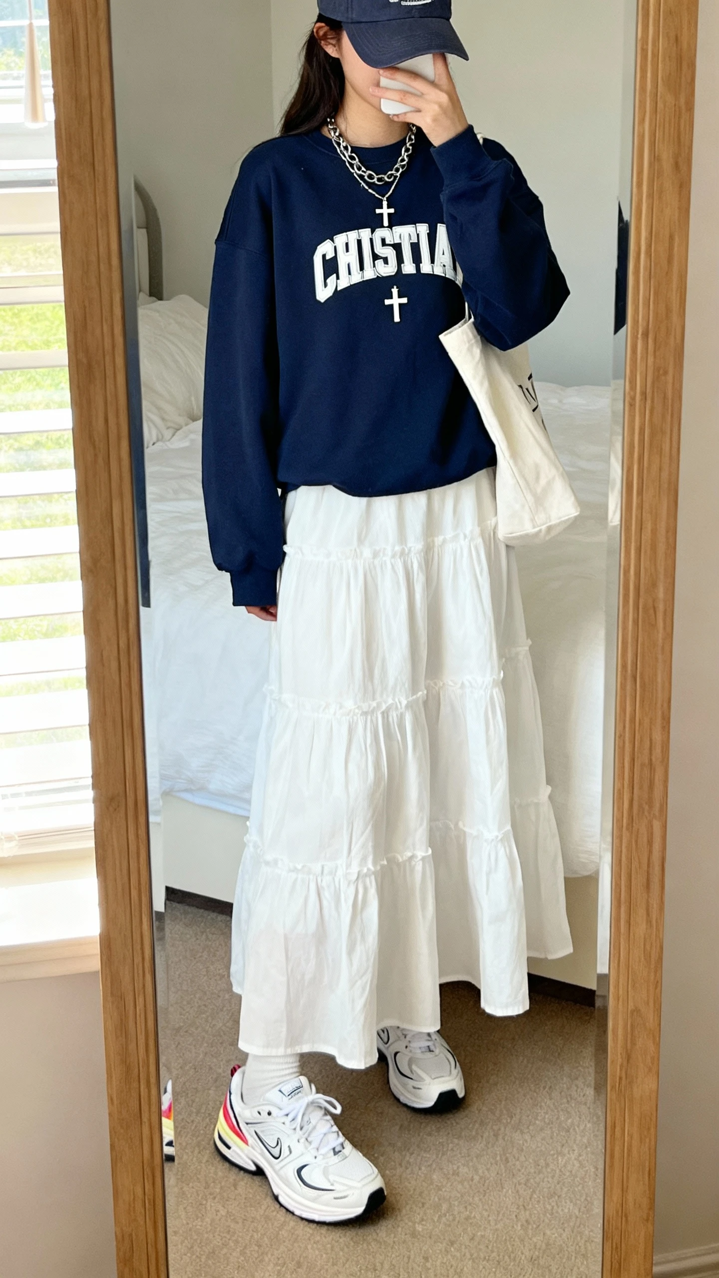 iPhone mirror selfie of a woman wearing a navy Christian crewneck with a white tiered poplin maxi dress, white retro runner sneakers with a subtle color pop, a baseball cap, layered chain necklace with a small cross charm, and a simple tote, face not visible, cropped at neck, bedroom mirror, bright morning light, iPhone photo quality.