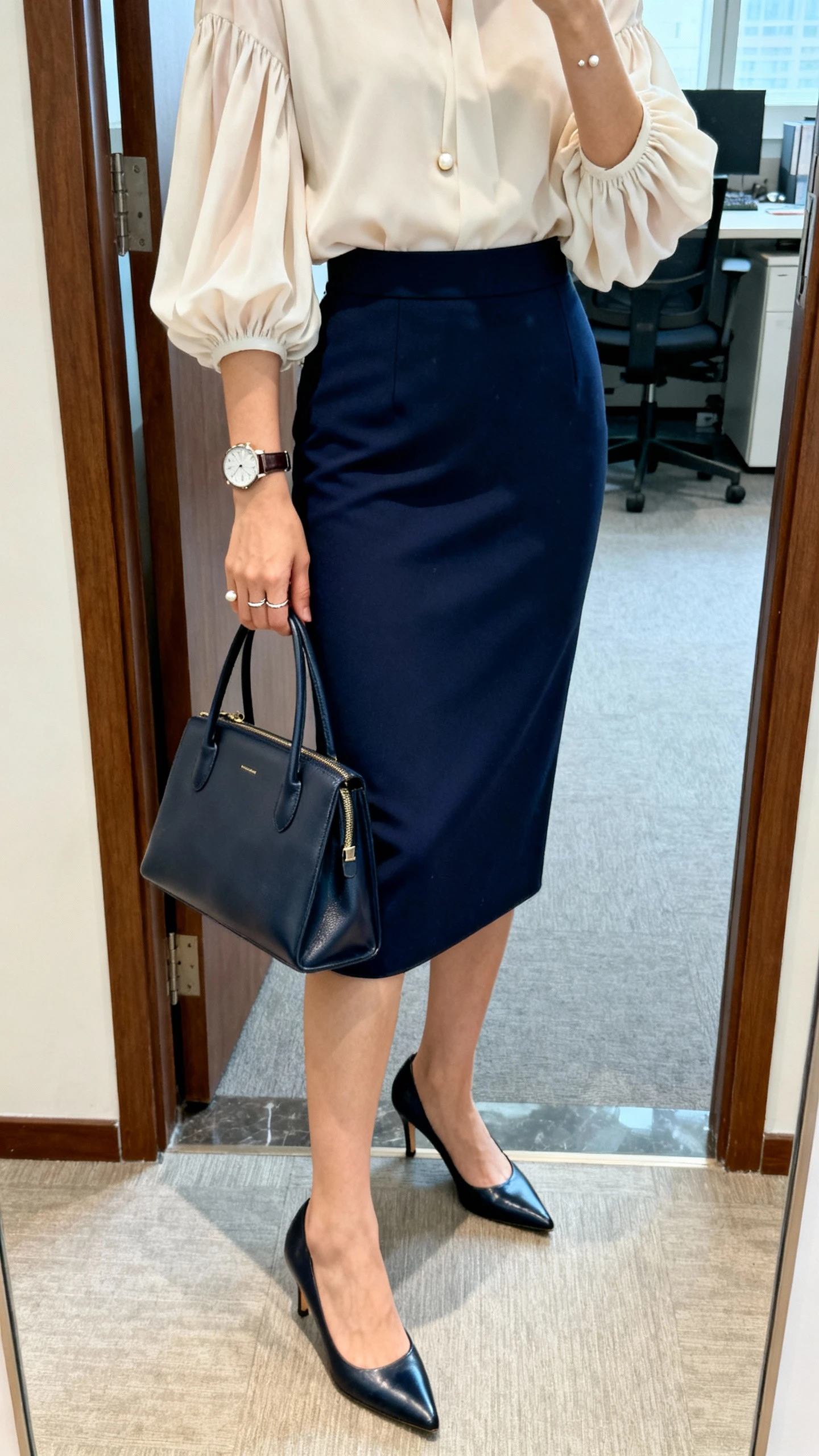 iPhone mirror selfie of a woman wearing a navy mid-calf high-waisted pencil skirt, flowy blouse with gathered sleeves, classic kitten heels, fine watch, delicate ring, small pearl studs, and a sleek tote; face not visible, cropped at shoulders, office entry mirror, bright indoor lighting, iPhone photo quality.