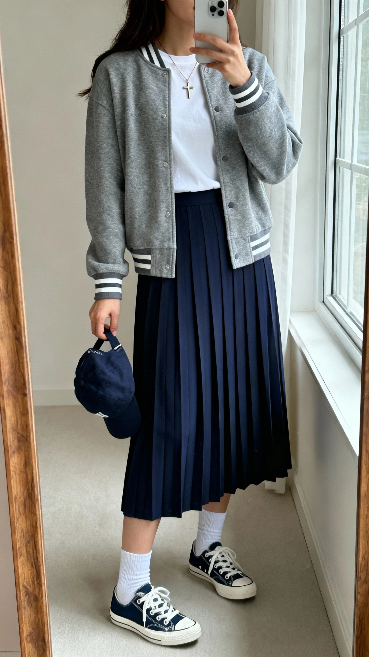 iPhone mirror selfie of a woman wearing a navy pleated midi skirt with a gray varsity cardigan layered over a modest white crew tee, white socks, retro navy-and-white sneakers, baseball cap in hand, and a delicate cross necklace; face not visible, side angle, full-length mirror, natural window light, iPhone photo quality.