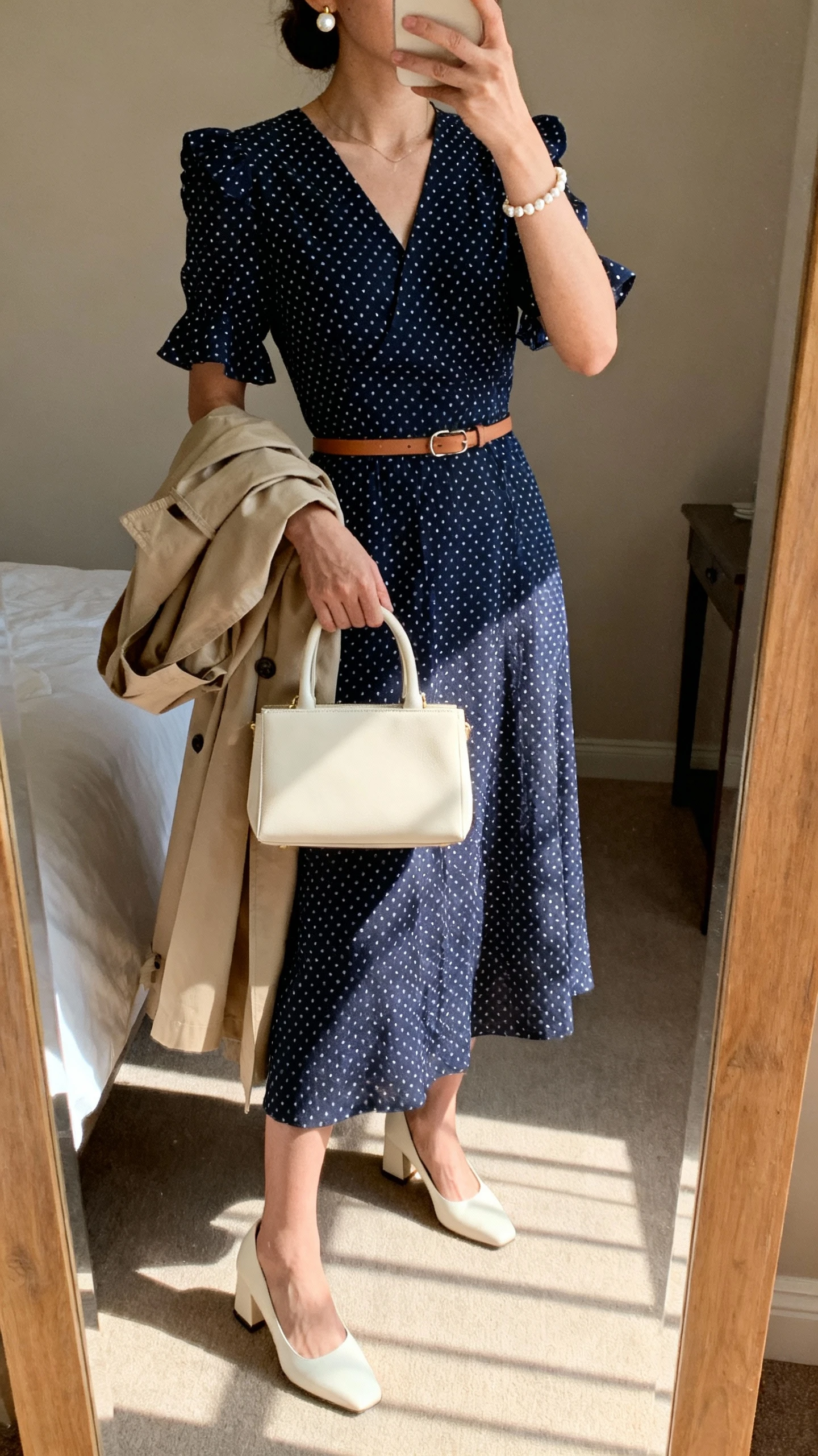 iPhone mirror selfie of a woman wearing a navy polka dot midi dress with a modest V-neck and flutter sleeves, cream block-heel pumps, a structured cream top-handle handbag, pearl stud earrings, a delicate pearl bracelet, a thin leather belt, and a beige lightweight trench draped over the arm, face not visible, cropped at neck, bedroom mirror, morning window light, iPhone photo quality.
