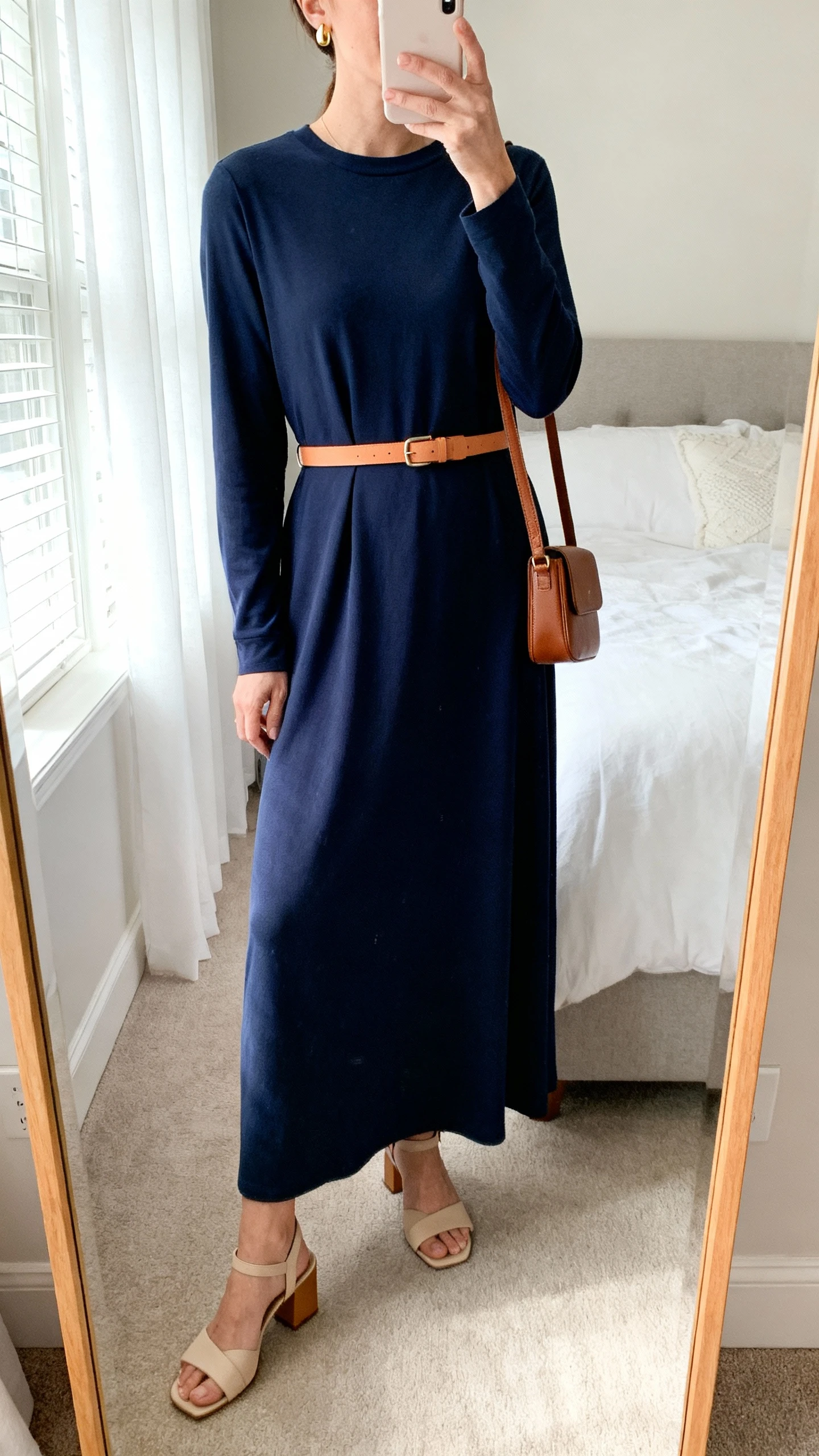 iPhone mirror selfie of a woman wearing a navy solid-color maxi dress with a modest crew neckline and long sleeves, paired with a thin tan leather belt, low beige-avoid note: tan block-heel sandals, a small structured brown crossbody, and simple gold stud earrings, face not visible, cropped at neck, bedroom mirror, morning window light, iPhone photo quality.