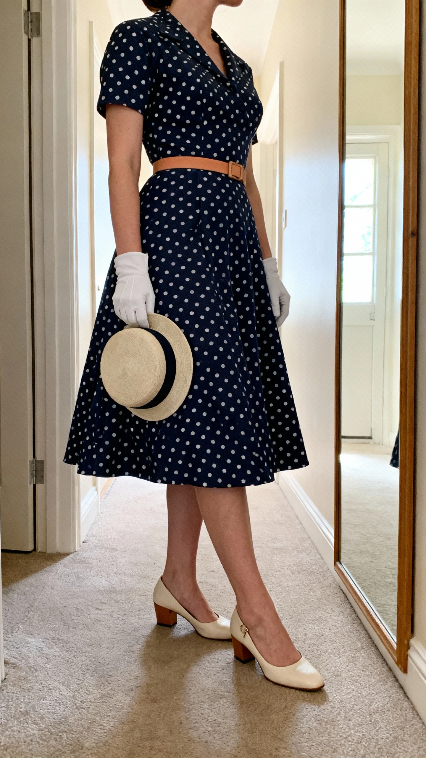 iPhone mirror selfie of a woman wearing a navy-with-white polka-dot fit-and-flare dress, thin camel belt, low block heels (slingback style), short white gloves, and a vintage-inspired pillbox hat in hand, face not visible, side angle in a hallway full-length mirror, morning indoor light, iPhone photo quality.