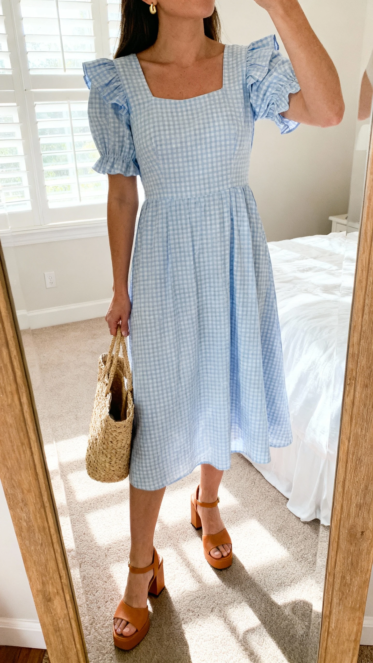 iPhone mirror selfie of a woman wearing a pastel-blue gingham garden-party midi dress with a modest neckline, flutter sleeves, and a gentle A-line, paired with tan block-heel sandals, a straw tote, and delicate gold studs, face not visible, cropped at neck, bedroom mirror, soft morning window light, iPhone photo quality.