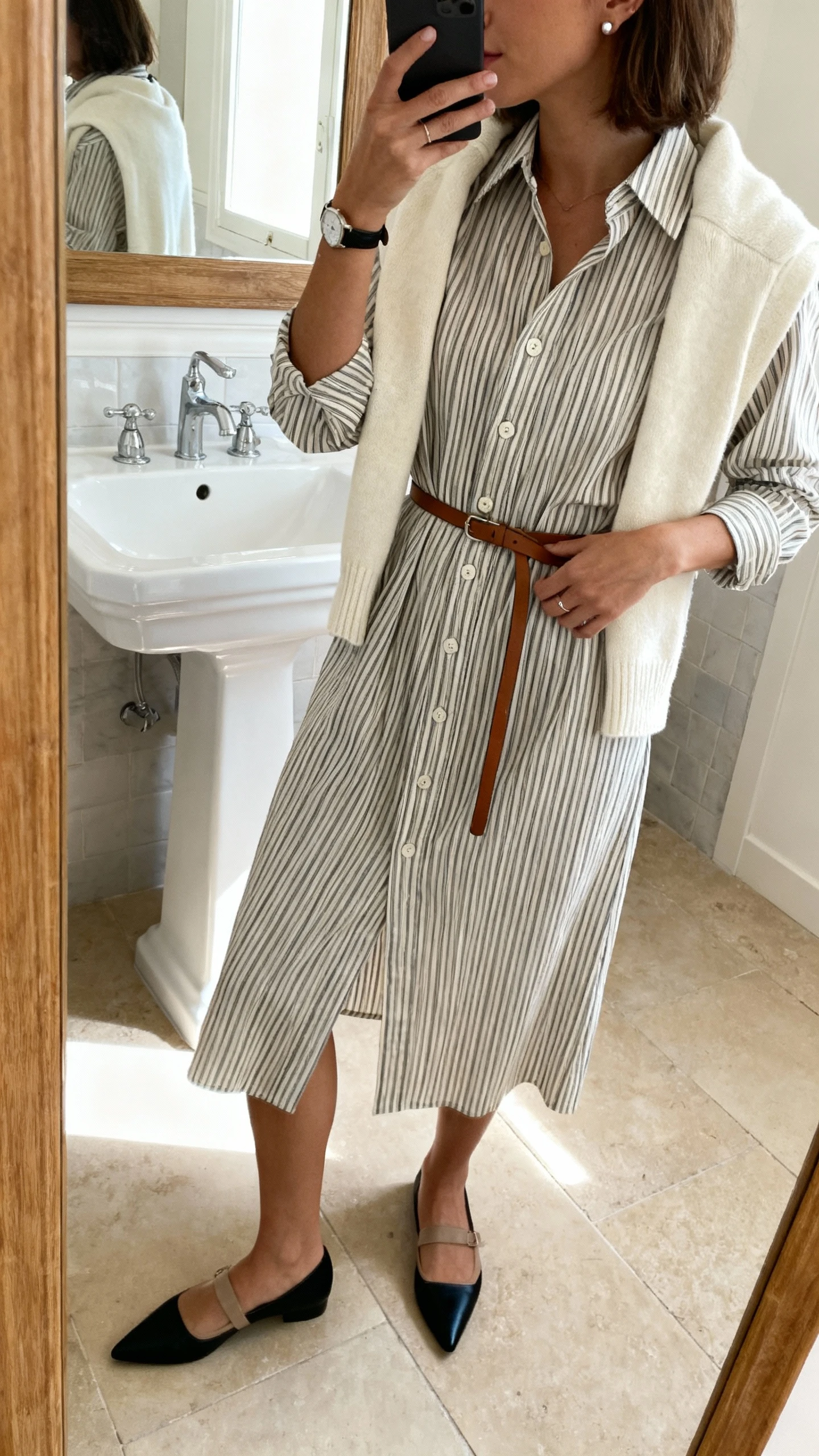 iPhone mirror selfie of a woman wearing a pinstripe shirt-dress with a belted waist and button-front, optional cream knit vest, pointed flats or low loafers in black or taupe, thin leather belt if needed, minimal watch and small stud earrings; face not visible, side angle in bathroom mirror, indoor natural lighting, iPhone photo quality.