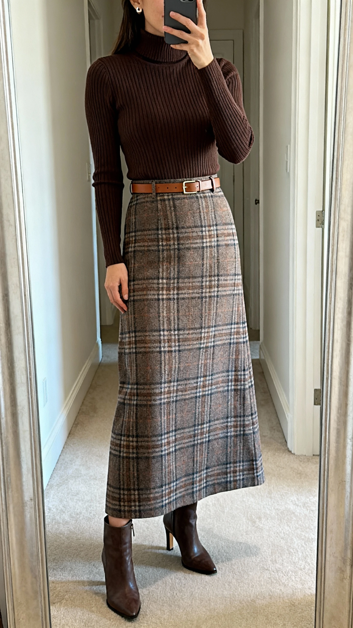 iPhone mirror selfie of a woman wearing a plaid maxi skirt, fitted ribbed turtleneck tucked in, waist cinched with a slim leather belt, heeled ankle boots, minimal stud earrings, face not visible, cropped at shoulders, full-length hallway mirror, natural daylight, iPhone photo quality.