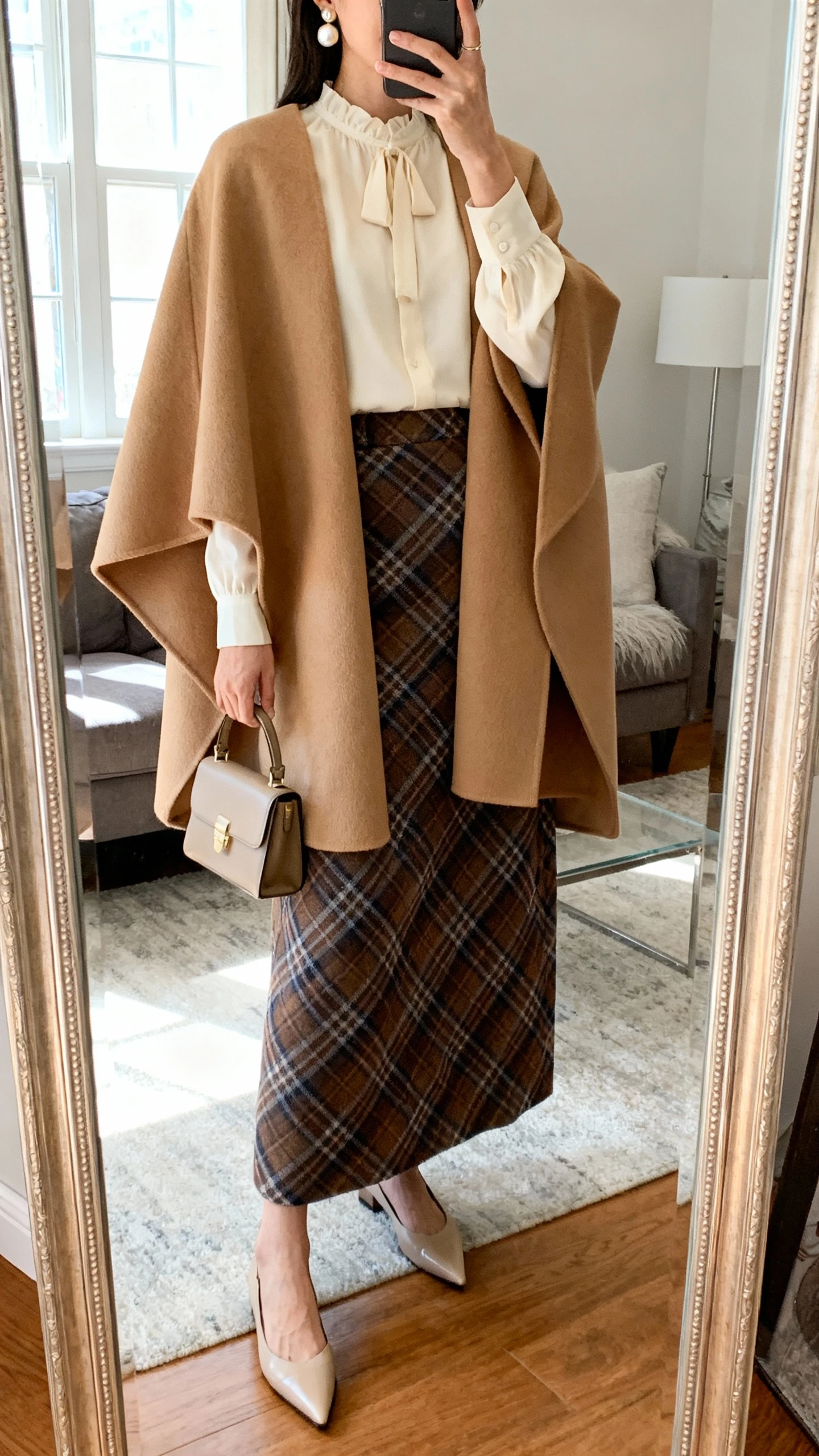 iPhone mirror selfie of a woman wearing a plaid maxi skirt, elegant camel cape coat draped over a soft ivory blouse, pointed-toe flats, small top-handle bag, pearl earrings, face not visible, cropped at neck, living room full-length mirror, natural window light, iPhone photo quality.