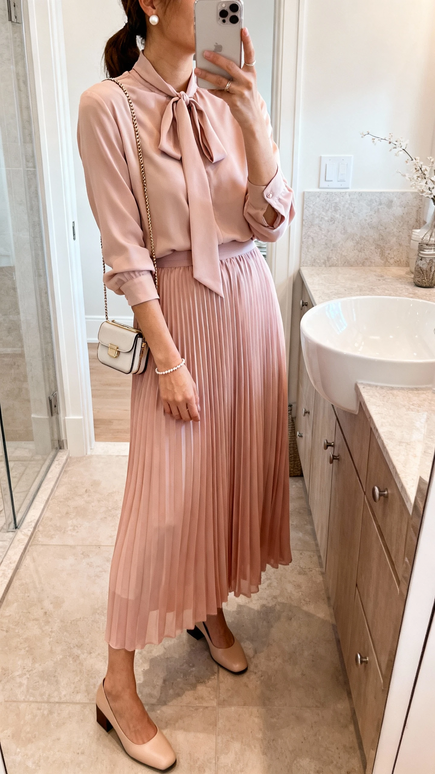 iPhone mirror selfie of a woman wearing a pleated chiffon maxi skirt in soft blush with a long-sleeve tie-neck blouse, low block heels, delicate bracelet, pearl studs, and a compact crossbody; face not visible, side angle, bathroom mirror, natural indoor lighting, iPhone photo quality.