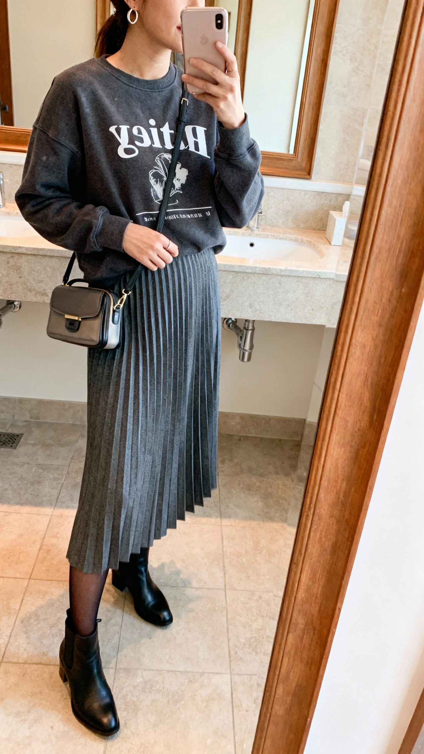 iPhone mirror selfie of a woman wearing a pleated midi skirt in charcoal with a graphic faith sweatshirt, black ankle boots, sheer black tights, small hoop earrings, compact shoulder bag; face not visible, side angle, bathroom mirror, warm indoor lighting, iPhone photo quality.