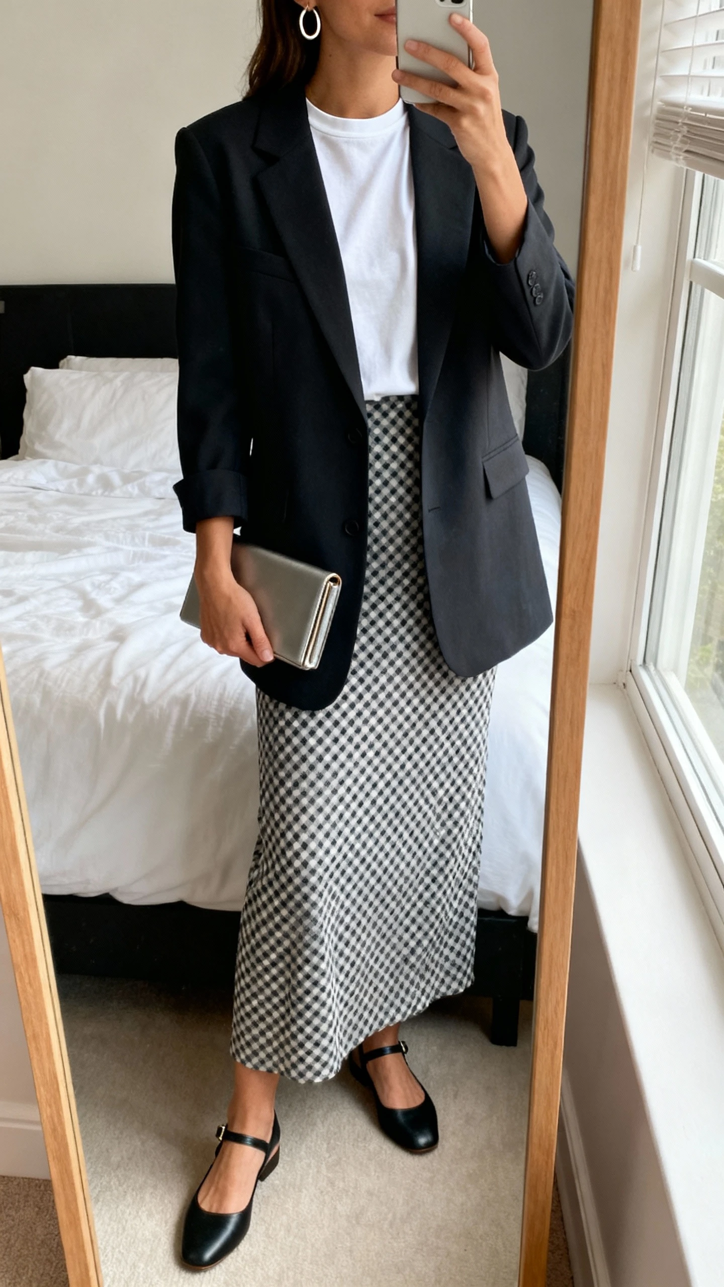 iPhone mirror selfie of a woman wearing a polished crewneck tee under a tailored blazer, gingham long skirt, ankle-strap flats, minimal hoop earrings, and a slim clutch; face not visible, cropped at shoulders, bedroom mirror, natural window light, iPhone photo quality.